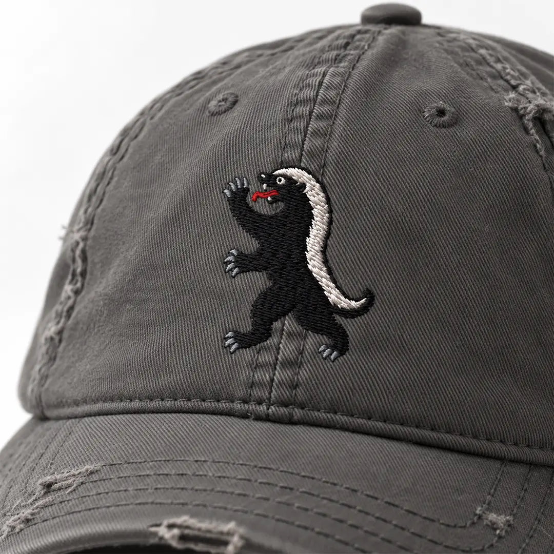 Close-up of the honey badger embroidery on a Charcoal Grey distressed cap, showing black fill stitches, pale back stripe, grey claws, and red tongue