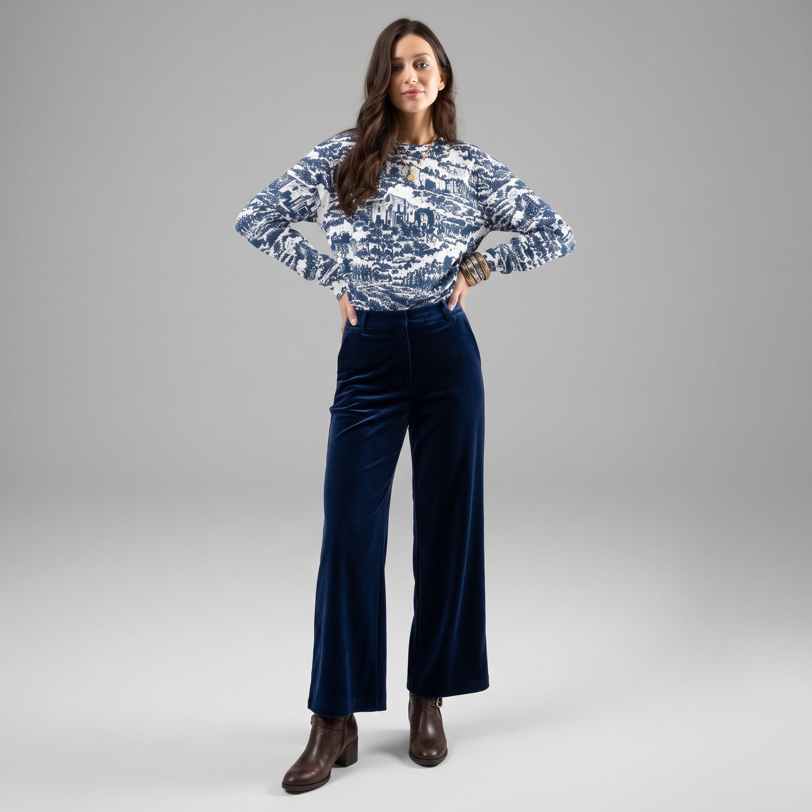 A woman modeling a long-sleeve patterned knit top and high-waisted navy blue velvet wide-leg pants, paired with brown leather ankle boots, against a neutral gray studio background.