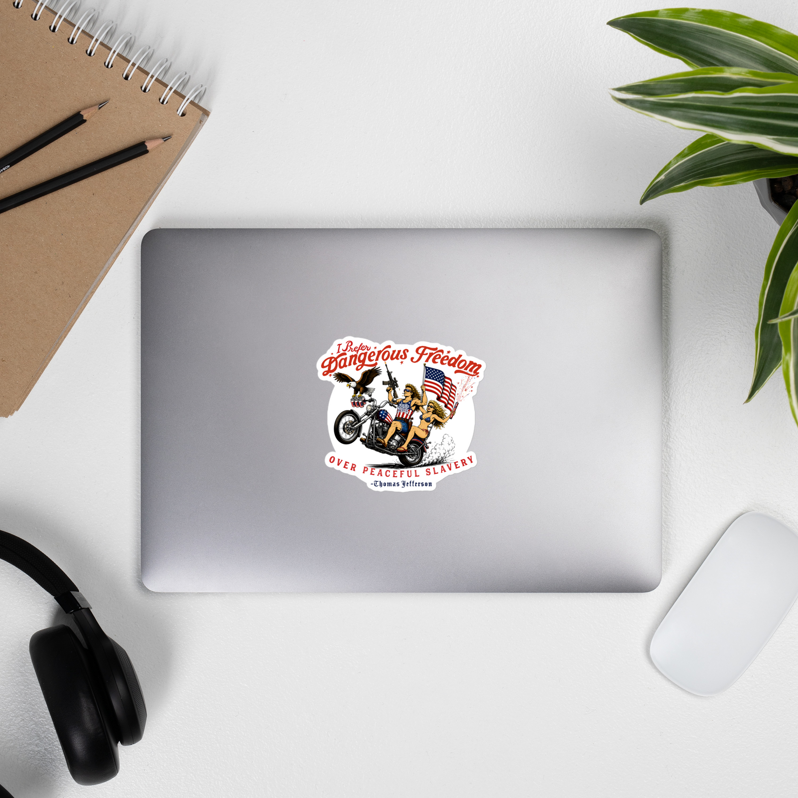 Laptop with a sticker on a desk with a notebook, pens, headphones, and a plant.