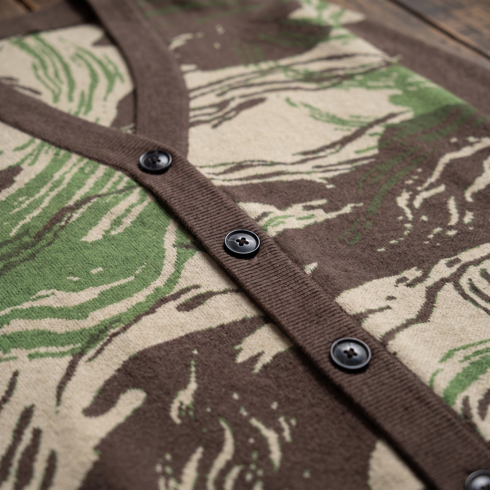 Close-up of a brown knit cardigan with a green and beige abstract camouflage-style pattern, featuring a ribbed brown V-neck trim and three dark buttons.