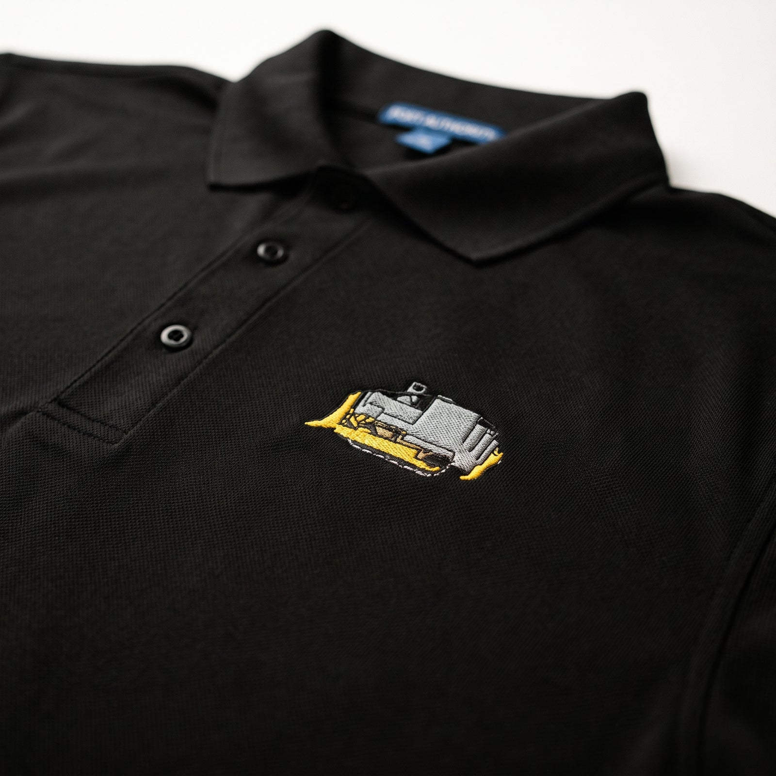 A close-up of a black polo shirt featuring a small embroidered killdozer graphic in gray and yellow on the left chest.