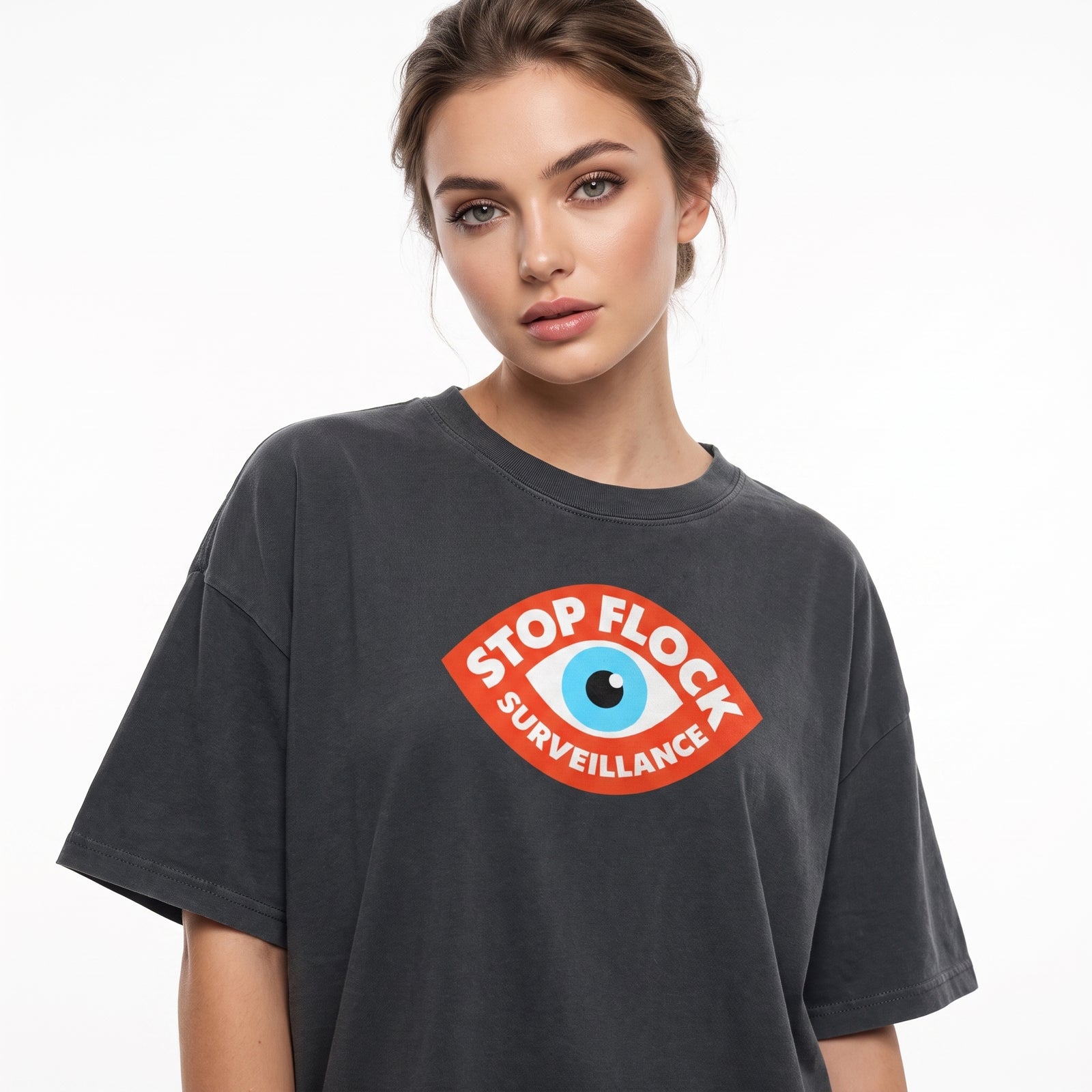 A woman wearing a dark gray washed t-shirt with a red, white, and blue graphic on the chest that reads 'STOP FLOCK SURVEILLANCE' around an eye illustration, set against a plain white background.