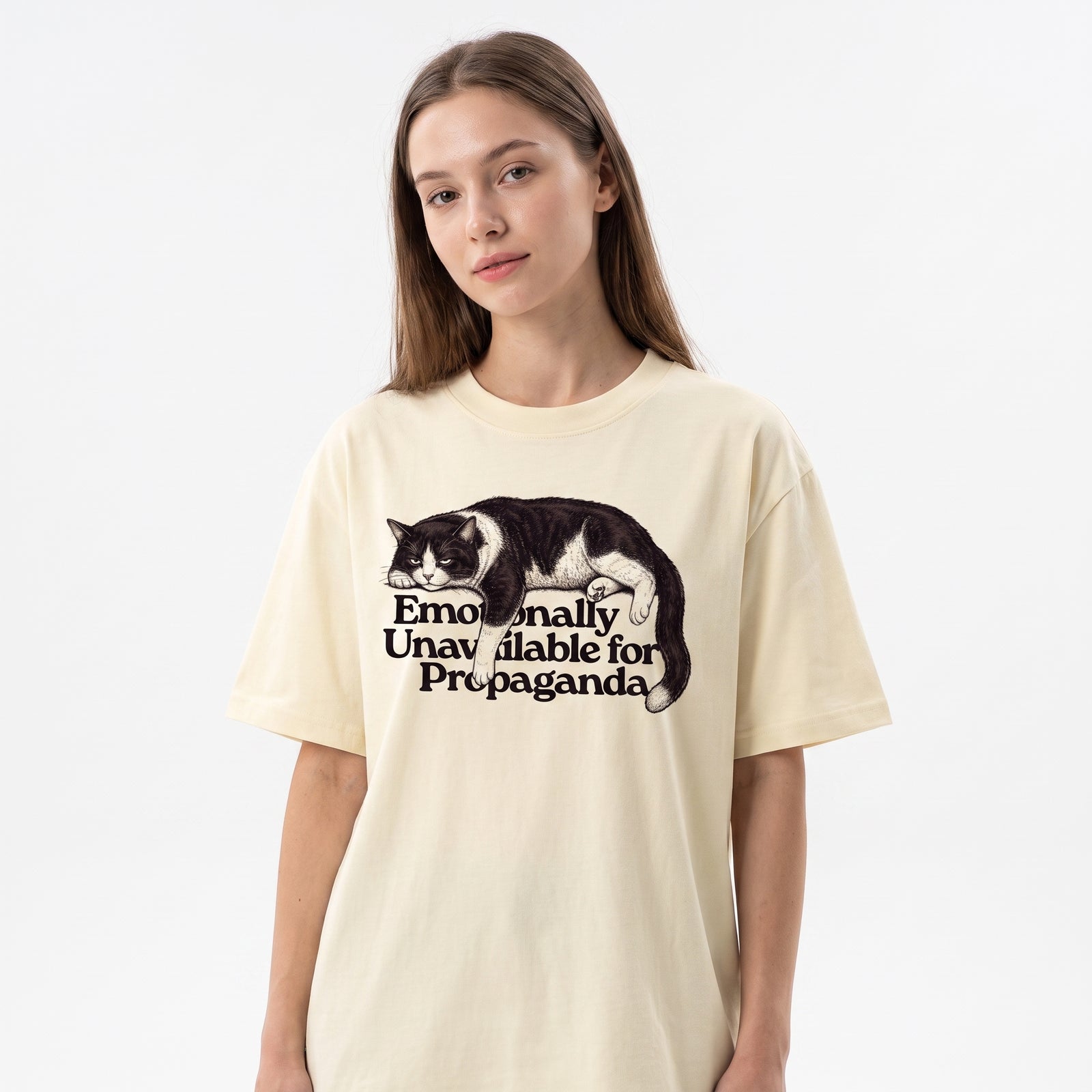 A woman wearing a cream-colored oversized t-shirt with a graphic of a sleeping black and white cat and the text 'Emotionally Unavailable for Propaganda'.
