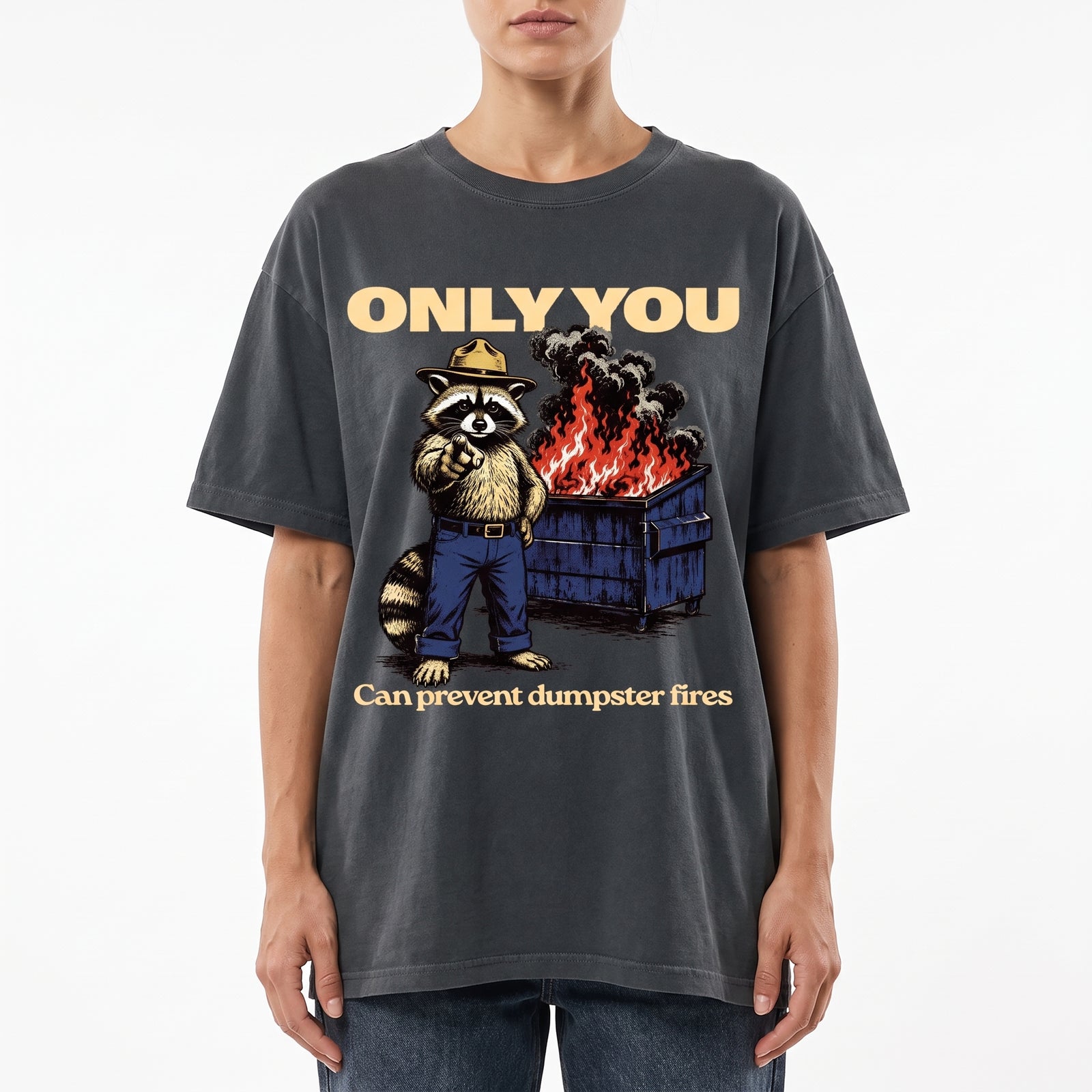 A model wearing a dark gray oversized graphic t-shirt. The shirt features a cartoon raccoon wearing a fire department hat standing next to a dumpster on fire, with the text 'ONLY YOU' above and 'Can prevent dumpster fires' below.