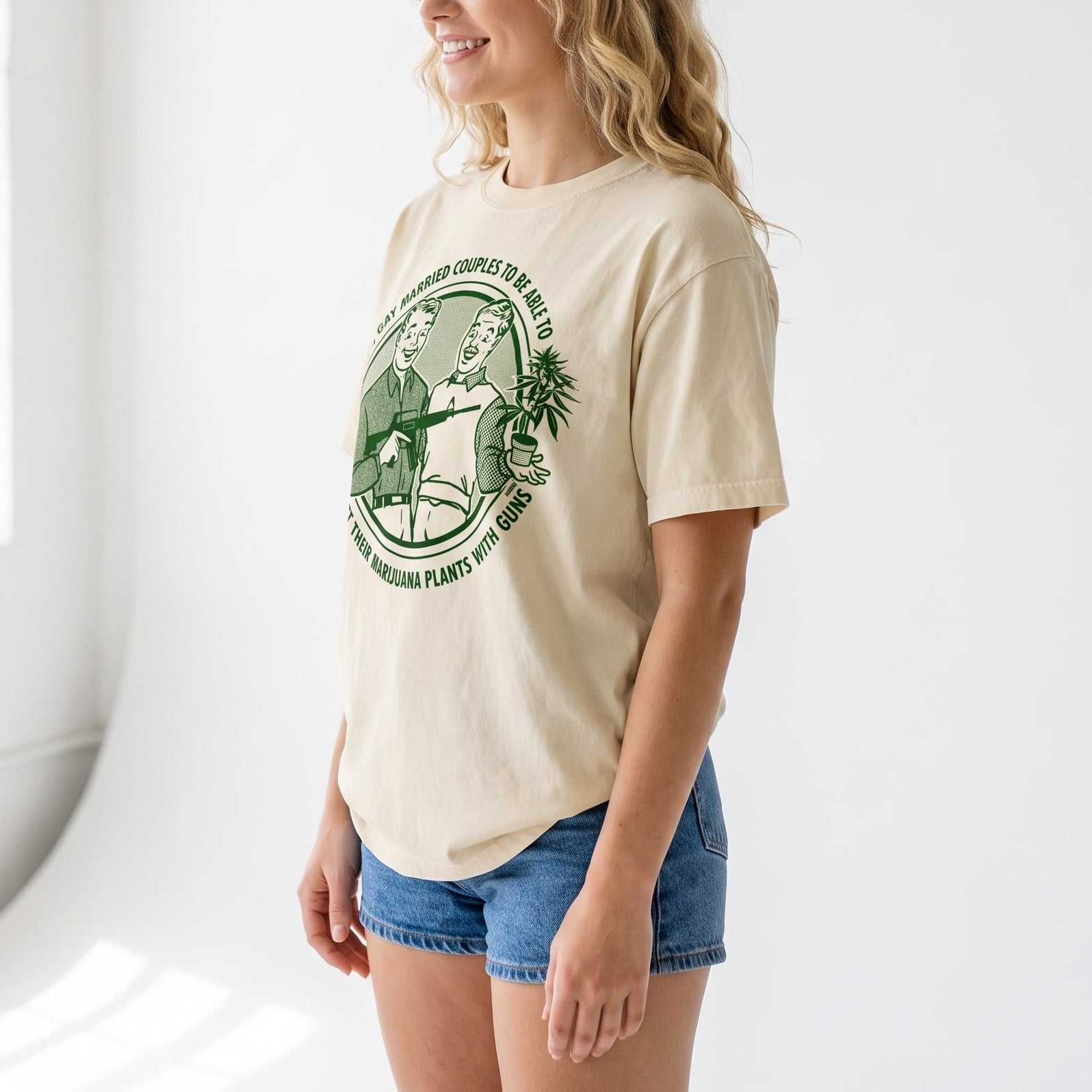 A woman with blonde wavy hair stands in a bright white studio setting, wearing a cream-colored t-shirt with a green circular graphic print and blue denim shorts.