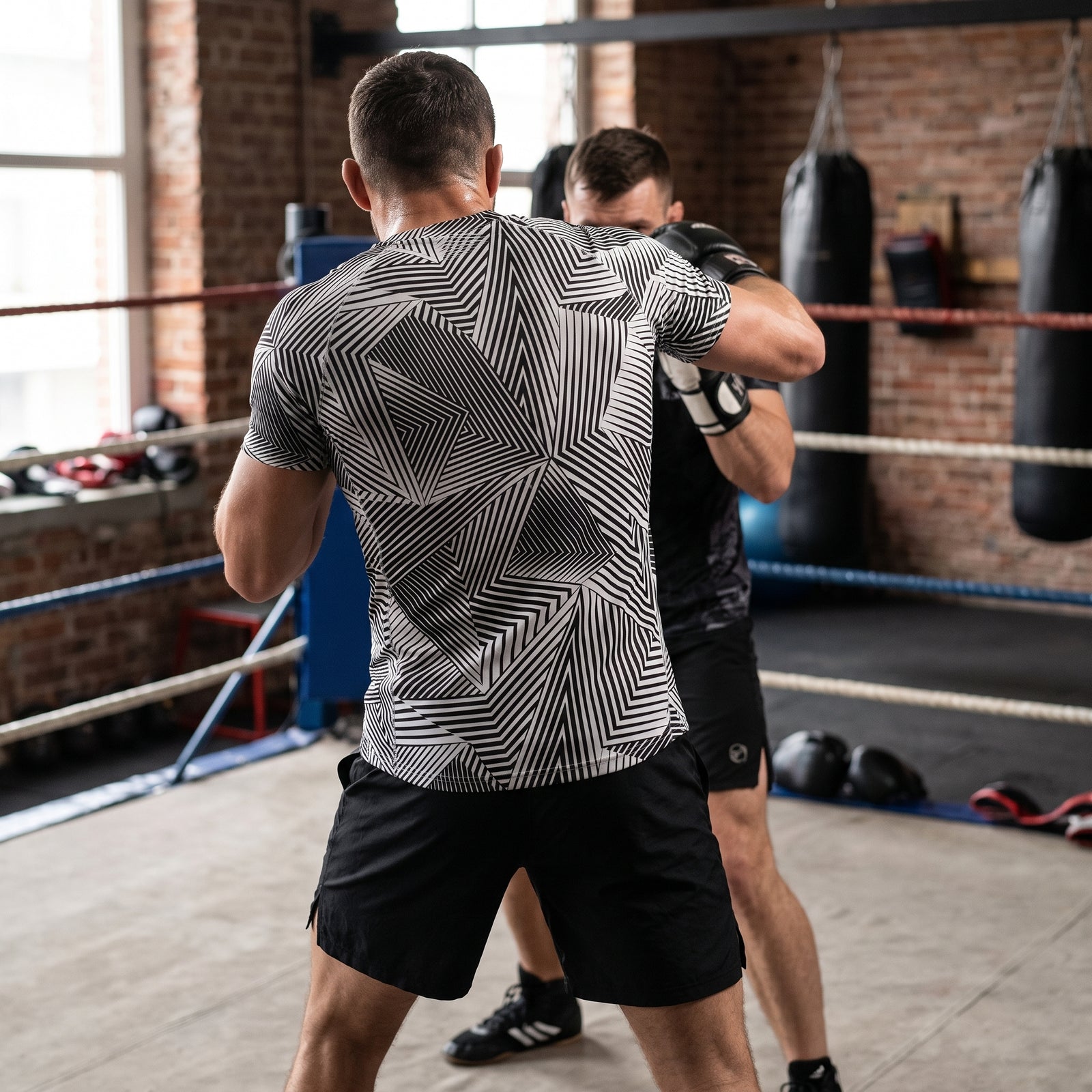 A man in a boxing ring wearing a black and white geometric patterned athletic t-shirt and black shorts, facing another boxer in a gym with brick walls and hanging punching bags.