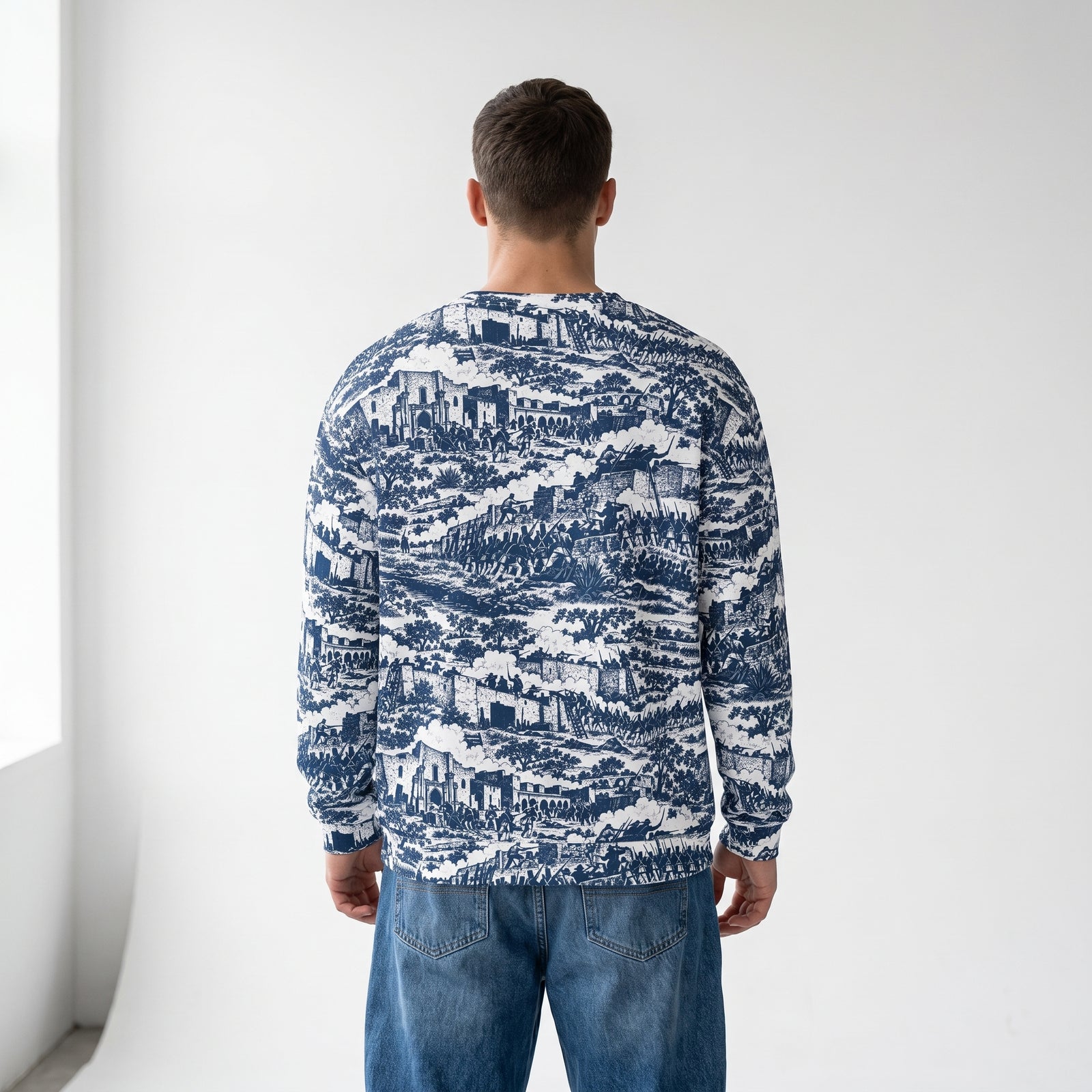 A man viewed from behind wearing a The Alamo long-sleeve crewneck sweater with a detailed blue and white landscape pattern, paired with blue jeans, set against a plain white studio background.