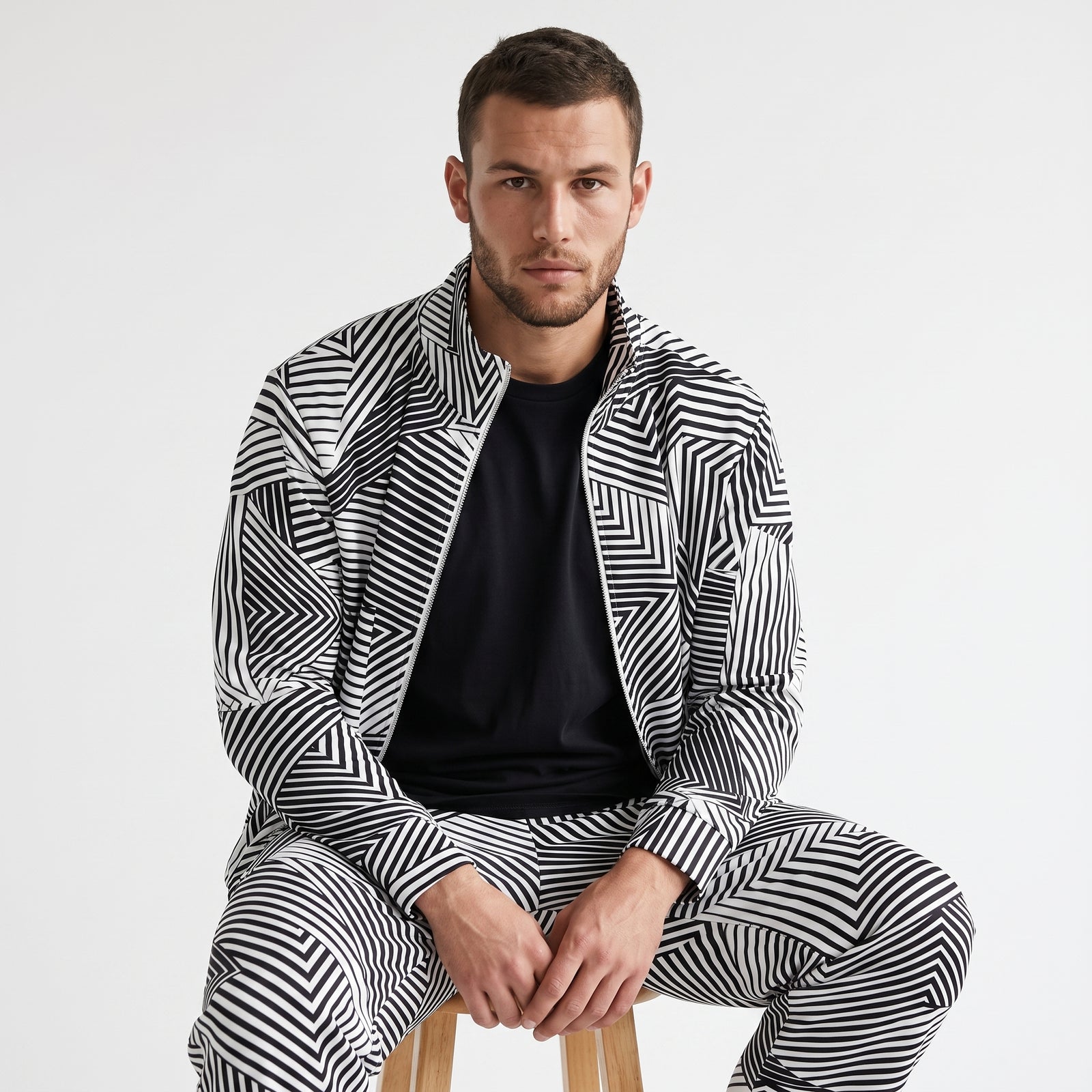 A man wearing a matching black and white geometric patterned zip-up jacket and trousers set, layered over a black t-shirt, sitting on a wooden stool against a plain white studio background.