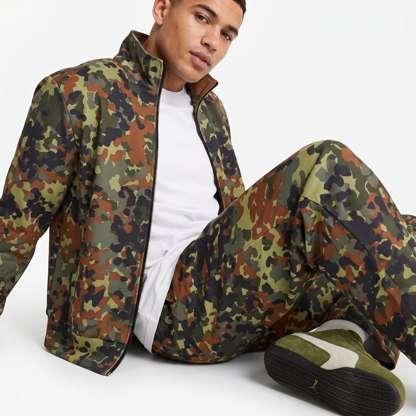 A man wearing a matching camouflage zip-up jacket and pants set, styled with a white t-shirt and olive green sneakers, sitting against a plain white studio background.