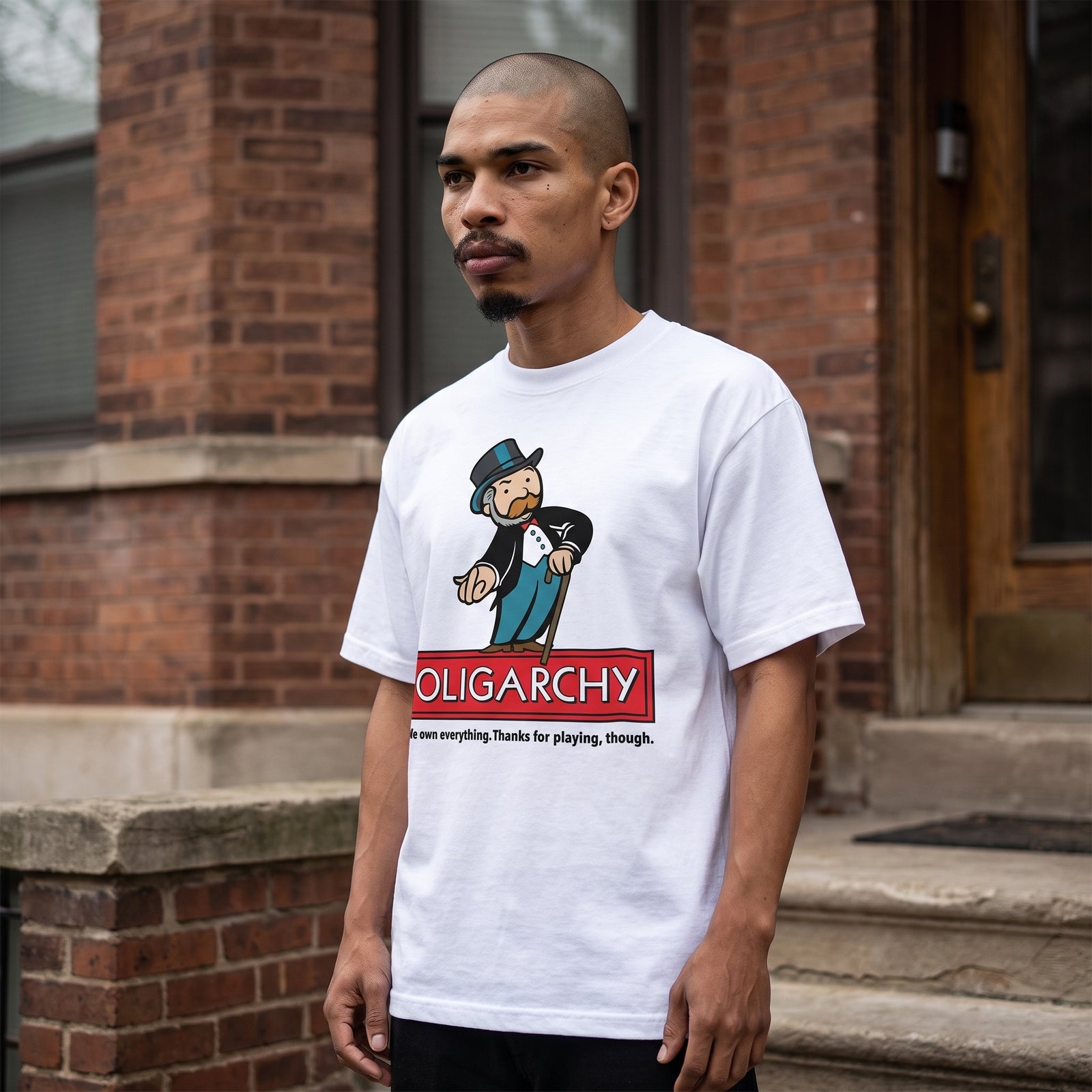 A man wearing a white graphic t-shirt with a cartoon character and the word 'OLIGARCHY' printed on the front, standing outdoors in front of a brick building.