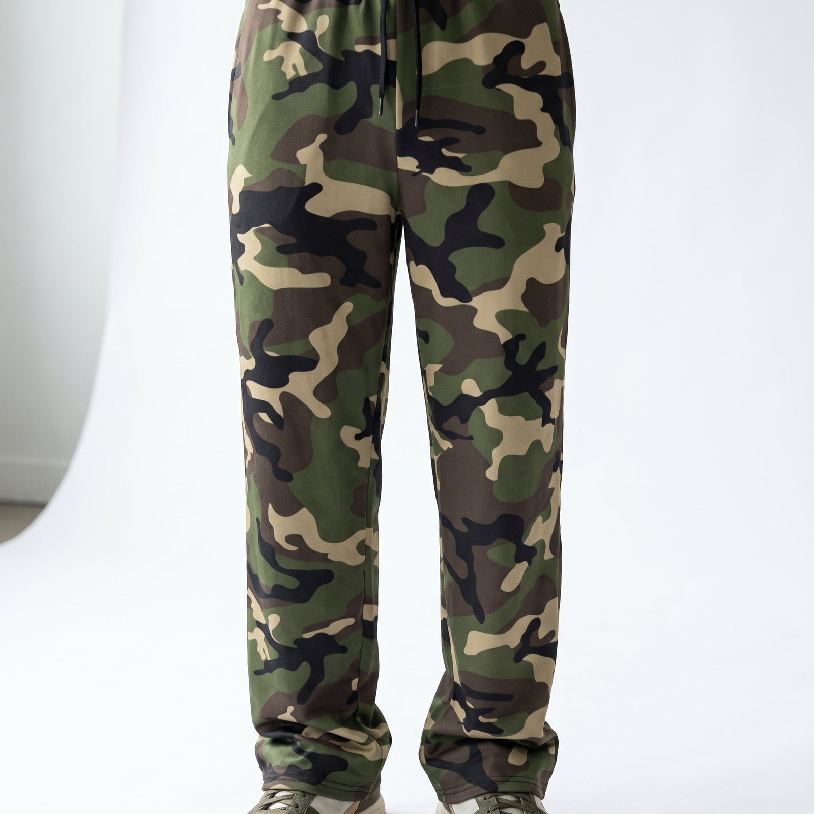A pair of camouflage sweatpants shown from the waist down, featuring a classic green, brown, black, and beige camo pattern. The pants have a drawstring waist and are paired with light-colored sneakers, photographed against a clean white studio background.