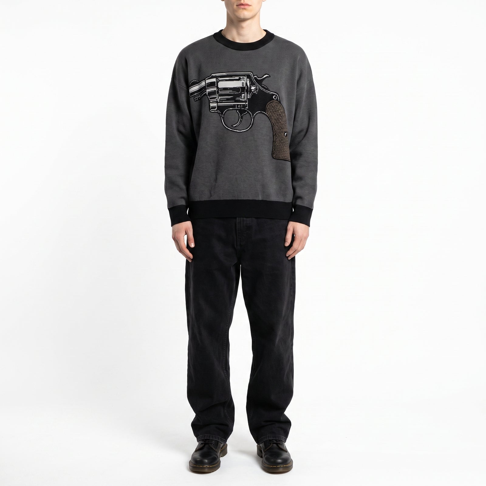 A model wearing a dark gray crewneck Jacquard-knit sweater with a large black and brown revolver graphic on the chest, paired with black pants and black shoes, shot against a plain white studio background.