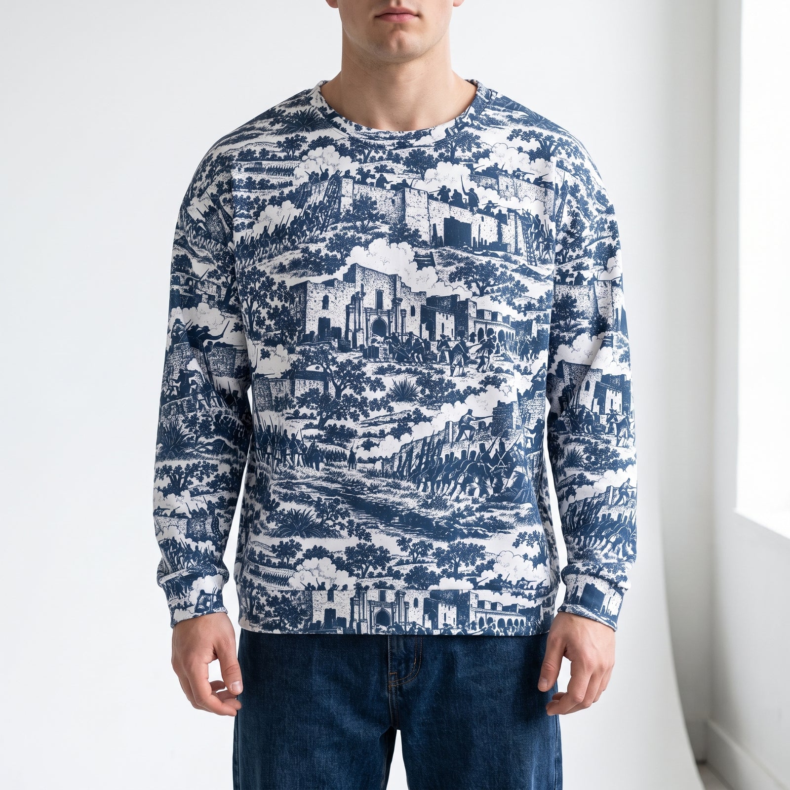 A man wearing a The Alamo Crewneck long-sleeve crewneck sweatshirt with a blue and white scenic landscape print, paired with dark blue jeans, standing against a white wall.