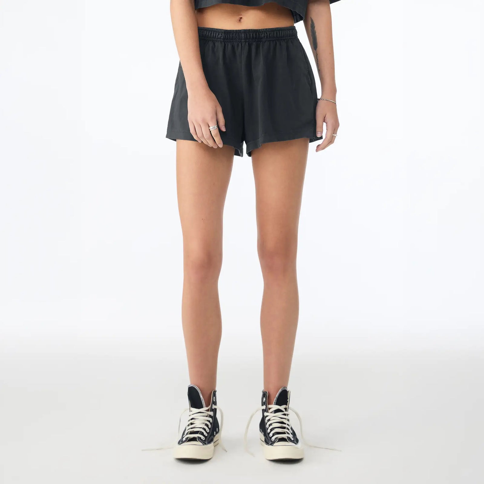 A model from the waist down wearing black athletic shorts, a dark gray cropped top, and black high-top sneakers with white laces, standing against a plain white studio background.