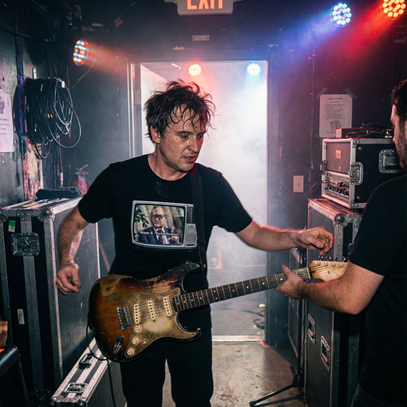 Man holding a guitar in a backstage area with stage lights and equipment.