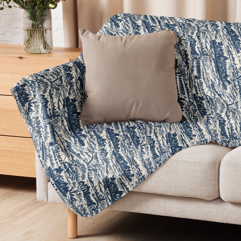 A blue and off-white patterned throw blanket draped over a light beige sofa, with a plain taupe pillow resting on top, in a softly lit living room setting.