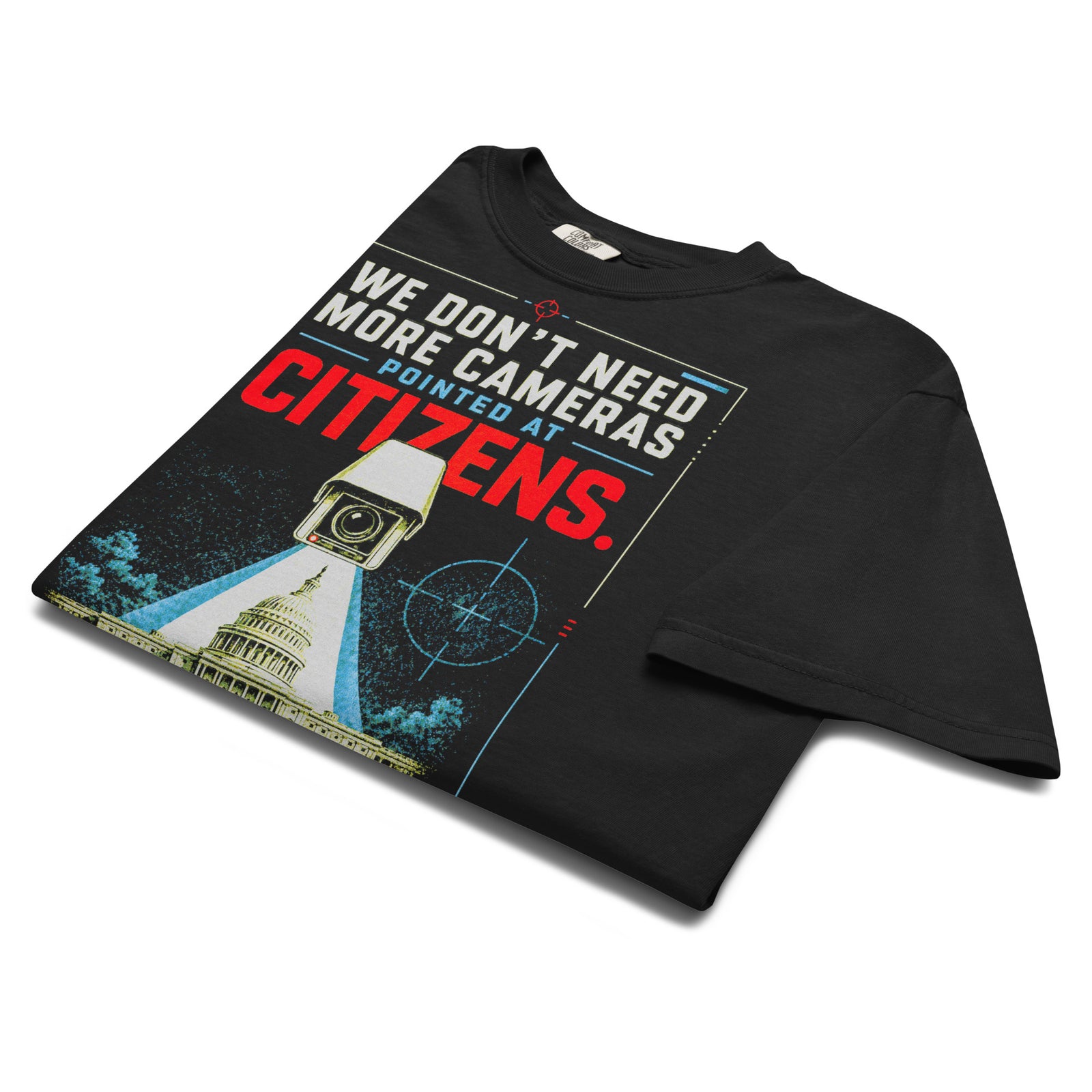 Cameras Pointed at Politicians Heavyweight Graphic Tee featuring a spy movie style surveillance camera aimed at the U.S. Capitol with bold red, cream, and blue typography on a black shirt.
