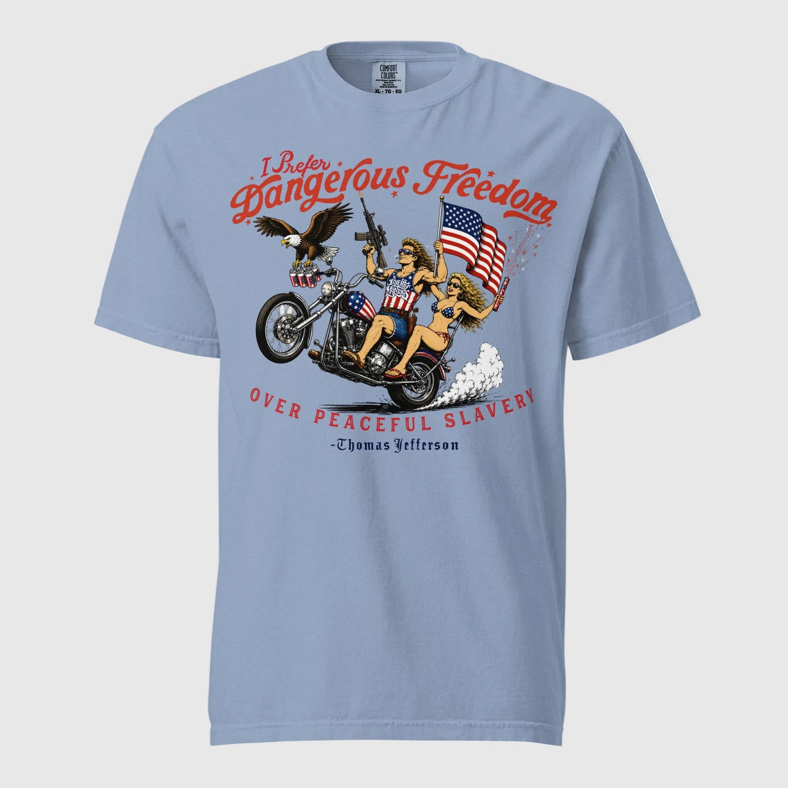 A light blue short-sleeve t-shirt featuring a retro-style illustration of a man and women on a motorcycle with American flags, an eagle, and fireworks. Red text above reads 'I Prefer Dangerous Freedom' and text below reads 'OVER PEACEFUL SLAVERY -Thomas Jefferson'. By Liberty Maniacs