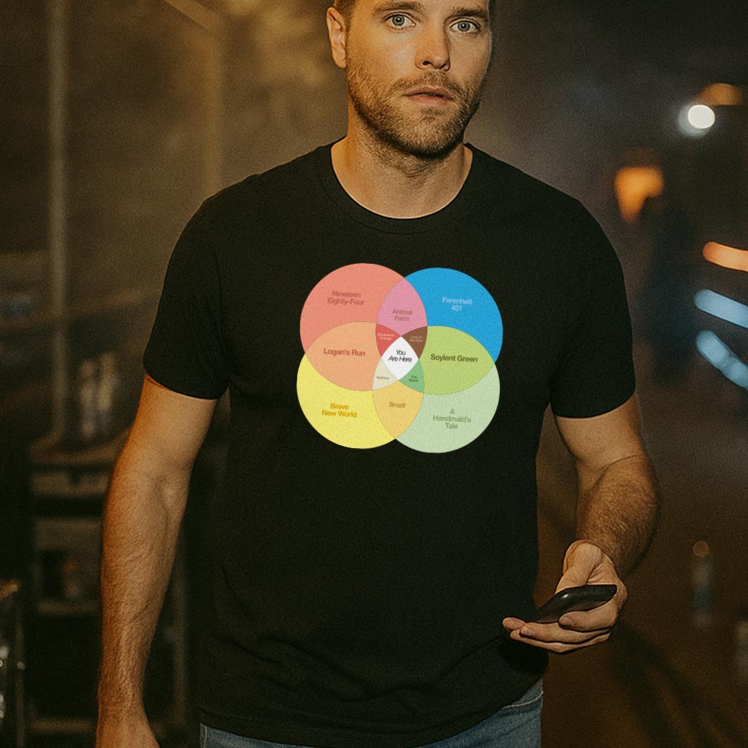 Man wearing a black t-shirt with a colorful Venn diagram design featuring dystopian fiction with the center reading "You Are Here", standing in a dimly lit room. The shirt is an exclusive from Liberty Maniacs.