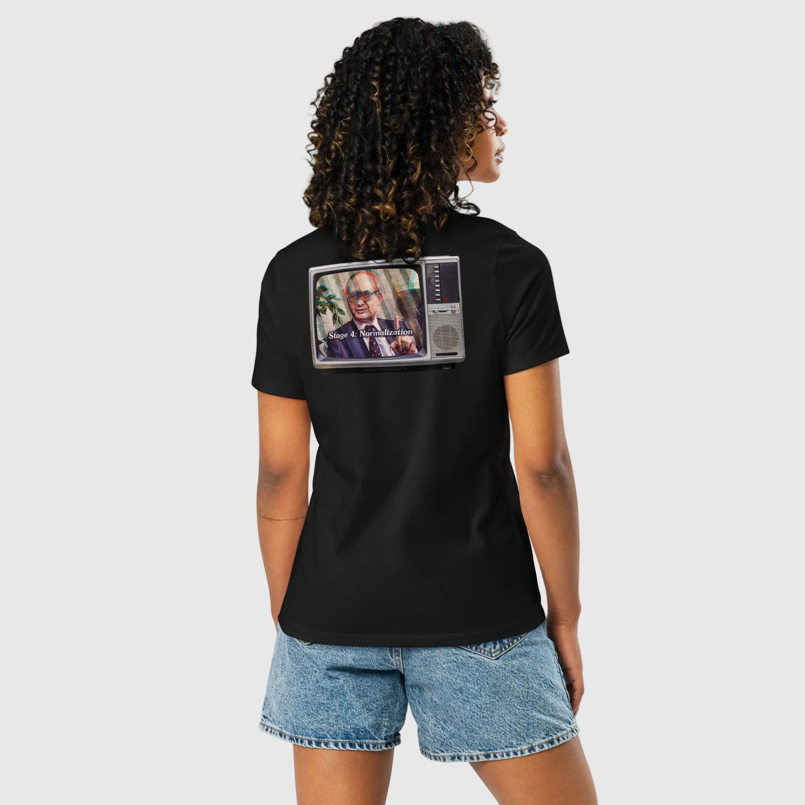 A woman shown from the back wearing a black short-sleeve t-shirt with a graphic print of a vintage television showing a man with sunglasses. She is also wearing denim shorts against a plain white background.