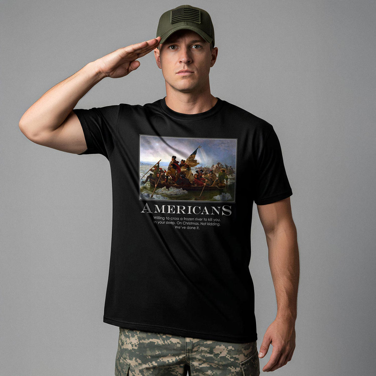 A man wearing an olive green tactical cap and camouflage pants stands at attention, saluting the viewer. He is wearing a black t-shirt from Liberty Maniacs. The t-shirt features a large graphic of the historical painting &quot;Washington Crossing the Delaware.&quot; Below the painting, the word &quot;AMERICANS&quot; is written in a large, bold font. Underneath, smaller text reads: &quot;Willing to cross a frozen river to kill you in your sleep. On Christmas. Not kidding. We&#39;ve done it.&quot;
