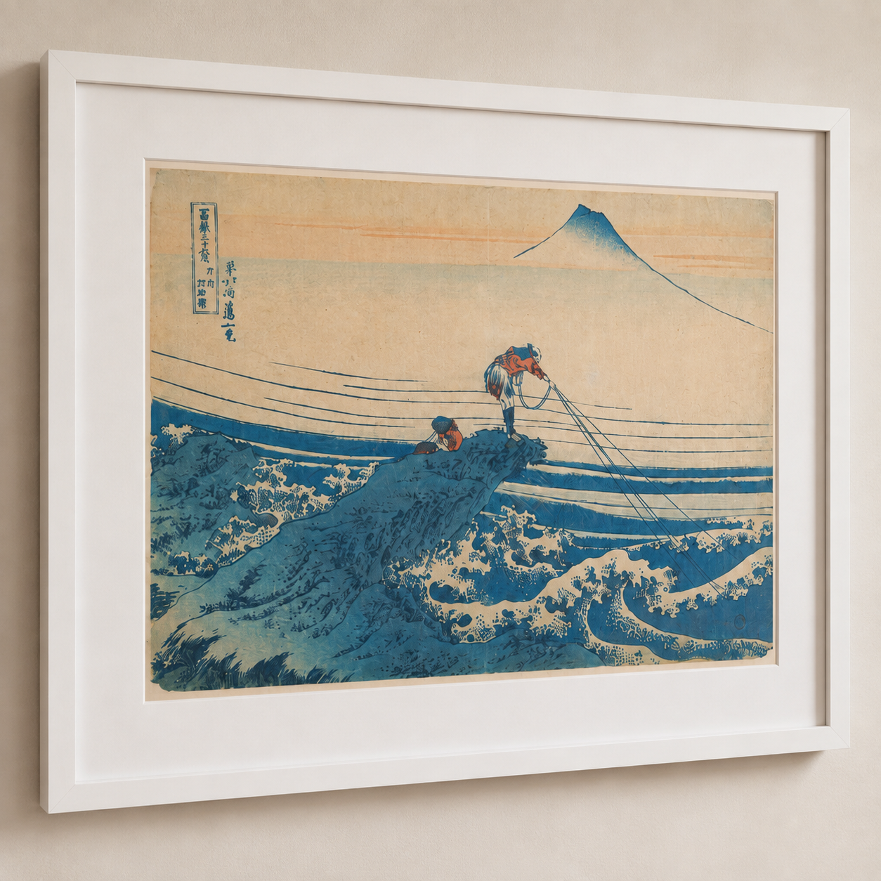 A framed art print of Hokusai's Great Wave off Kanagawa, featuring a large blue wave and Mount Fuji against a warm beige sky, presented in a white frame on a neutral beige wall.
