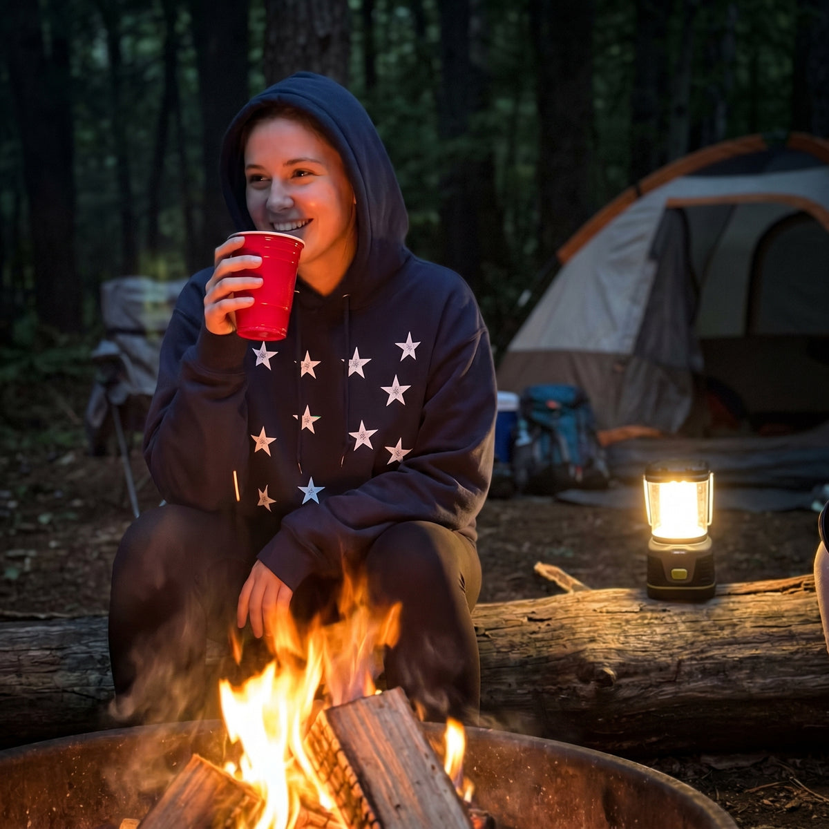 Woman sitting by a campfire wearing a Green Mountain Boys 13 Star Heritage Hoodie, holding a red cup, with a tent and lantern in the background.