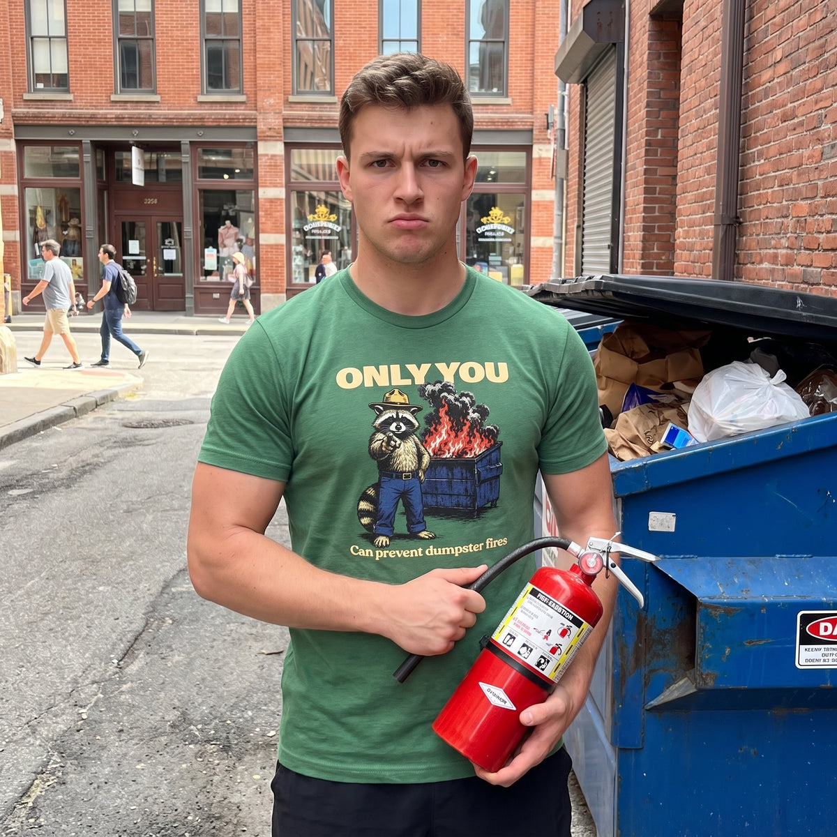 Man stands in an ally next to a dumpster holding a fire extinguisher, wearing a green t-shirt that says 'Only You Can Prevent Dumpster Fires" with a racoon park ranger on it.