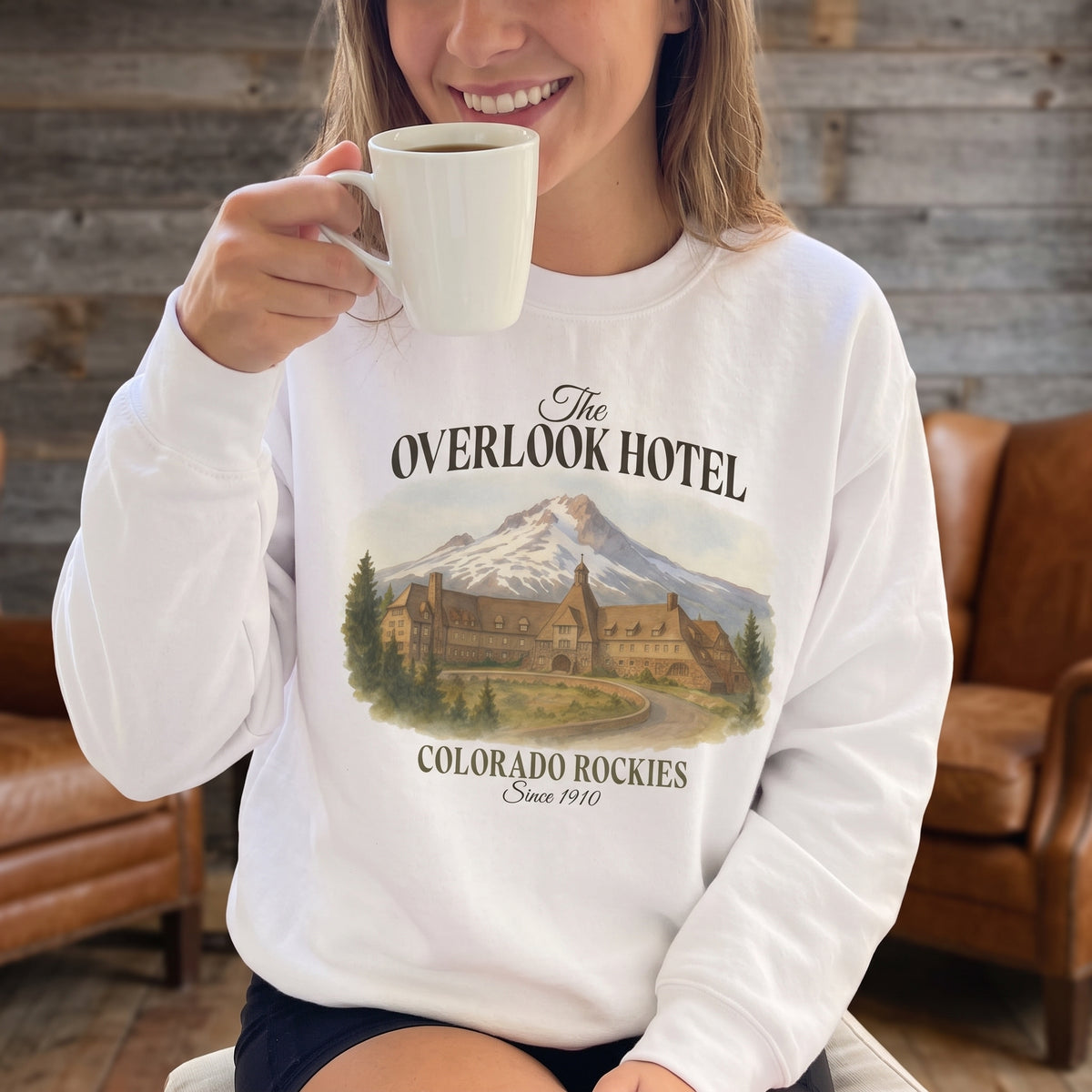 Woman wearing a white Overlook Hotel sweatshirt while holding a mug in a rustic lodge room