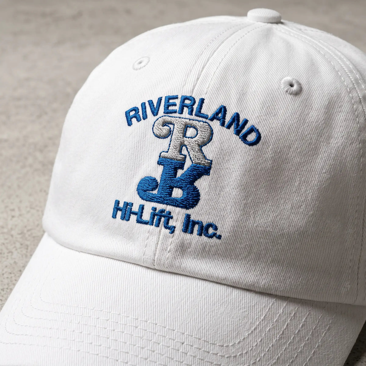 A white cotton baseball cap with blue and silver embroidered text reading 'RIVERLAND HB' and 'HI-Lift, Inc.' on the front panel, photographed close up on a neutral textured surface.