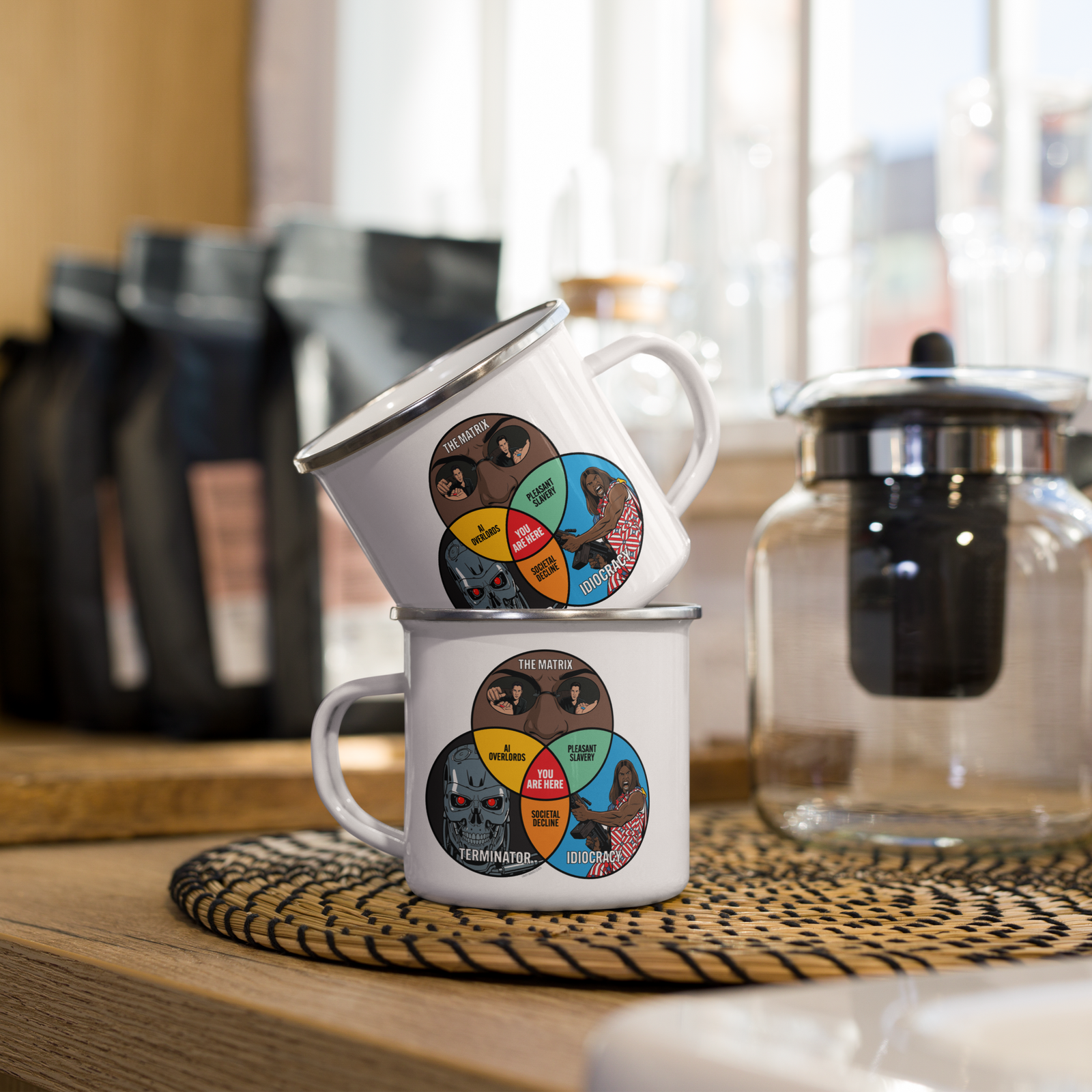 Two stacked white enamel camping mugs with a colorful circular graphic on the front, set on a wooden table next to a glass teapot and coffee bags in a warm, softly blurred kitchen setting.