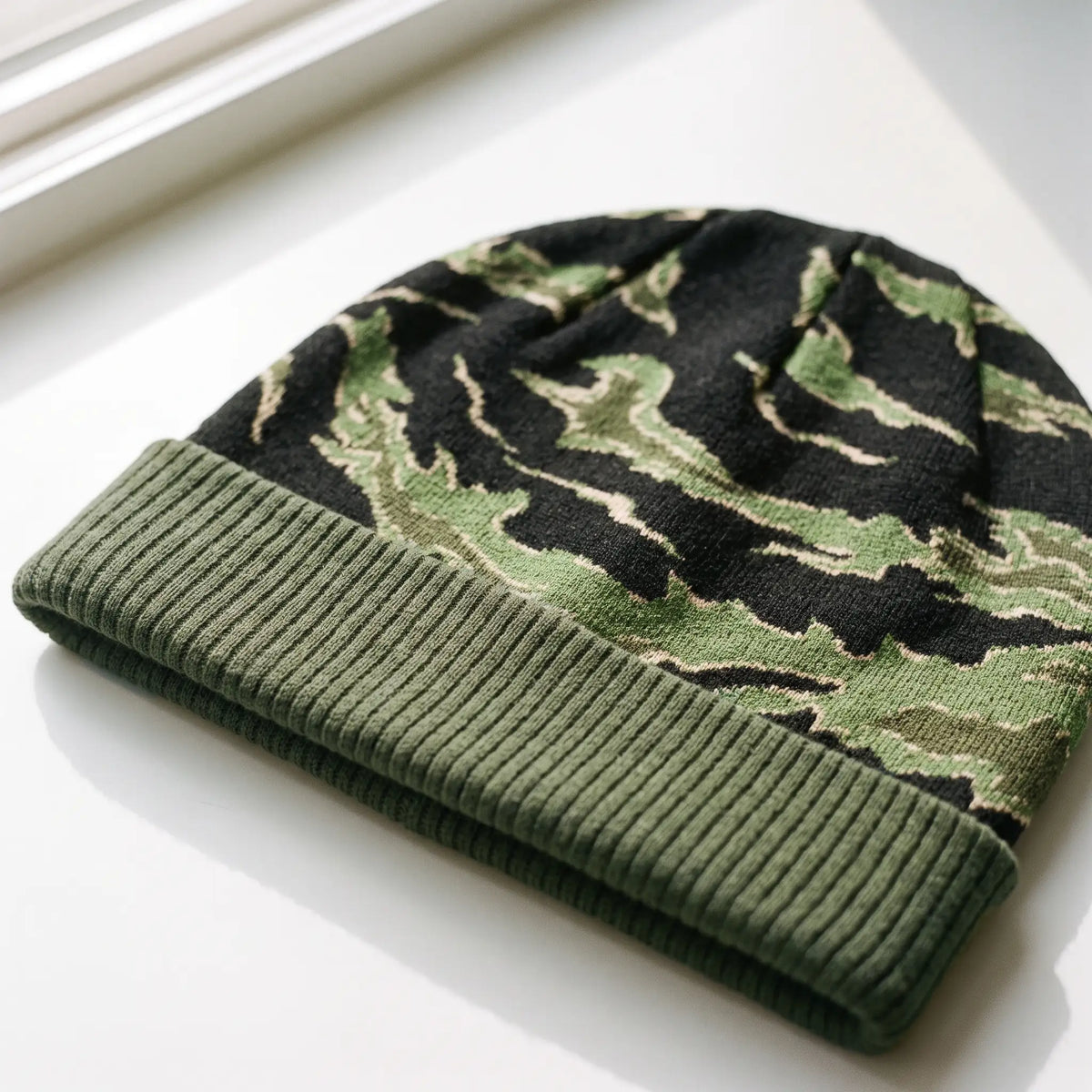 A knit beanie hat with a green and black camouflage pattern and a solid olive green ribbed cuff, resting on a white surface near a window.