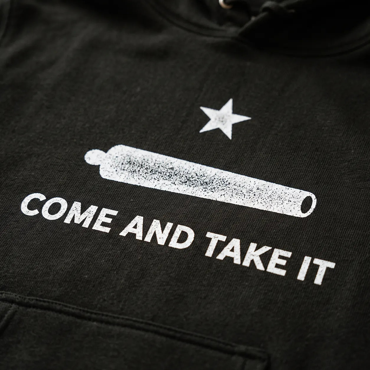 Close-up of a black hoodie featuring a white distressed graphic of a cannon, a star, and the text 'COME AND TAKE IT' on the chest.