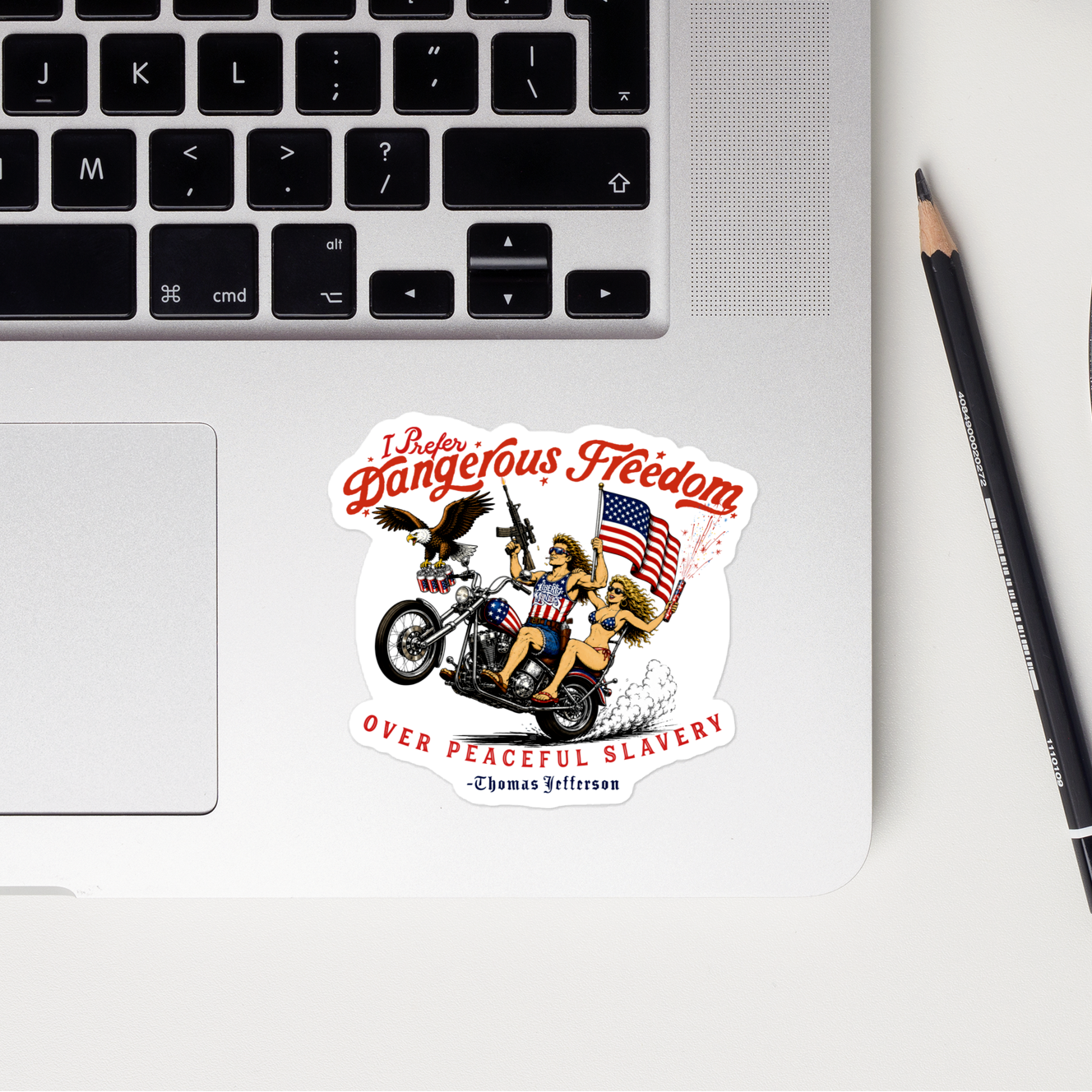 A circular sticker with a biker illustration and the text 'Prefer Dangerous Freedom over Peaceful Slavery' by Thomas Jefferson, shown on a silver laptop. The laptop is part of a styled flat-lay on a white surface alongside a black smartphone, a pencil, a blue pen, and a red notebook.