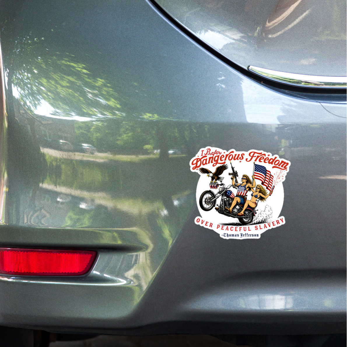 The Liberty Maniacs sticker of two riders on a motorcycle shooting off guns and fireworks as an eagle carries a sixpack of beer alongside their hog doing a wheely. The Thomas Jefferson quote "I Prefer Dangerous Freedom over Peaceful Slavery" surrounds the scene. The sticker is applied to the rear bumper of a gray car, parked outdoors on a paved surface.