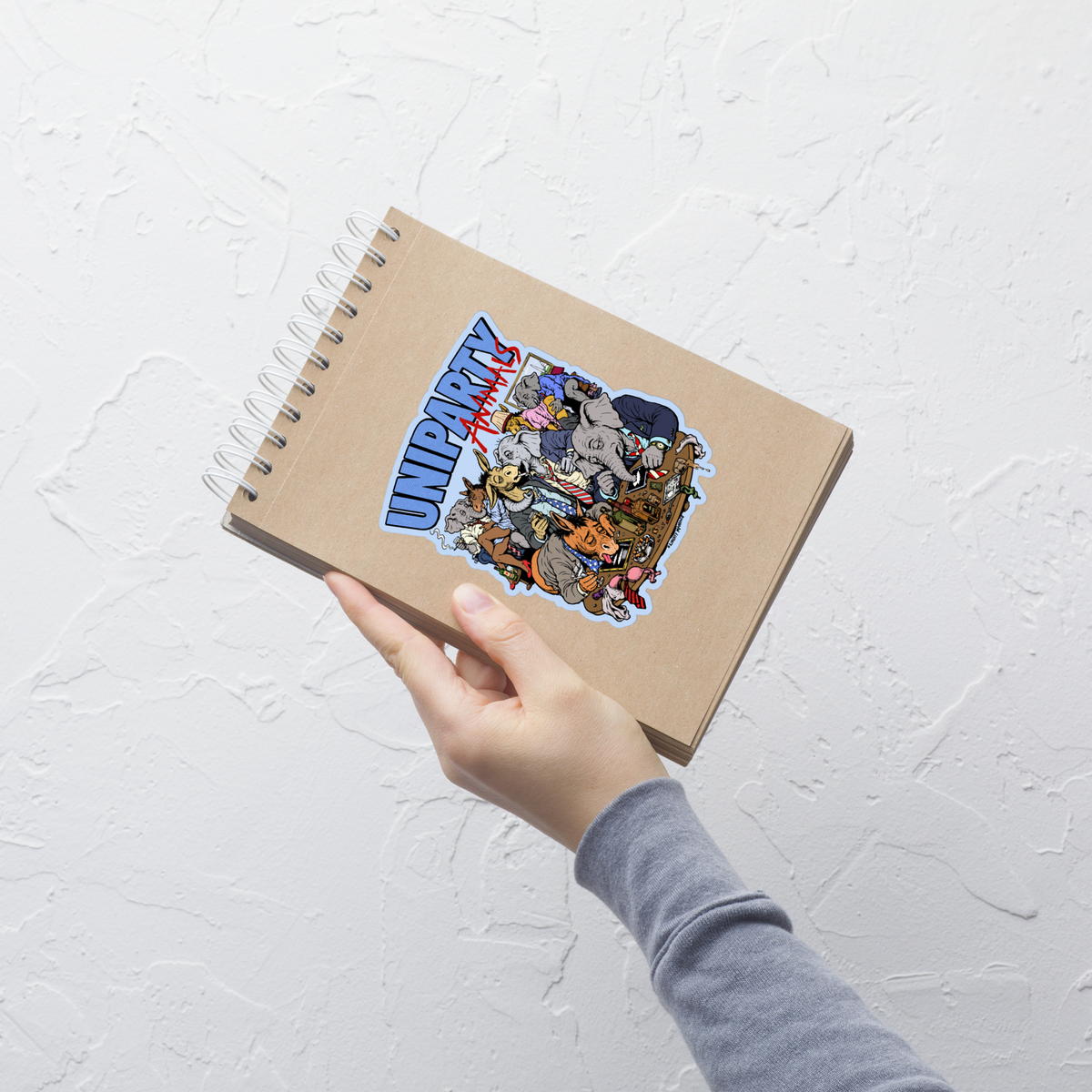 A hand holding a spiral-bound notebook with a colorful 'UNPARTY' sticker on the cover, set against a textured white wall.