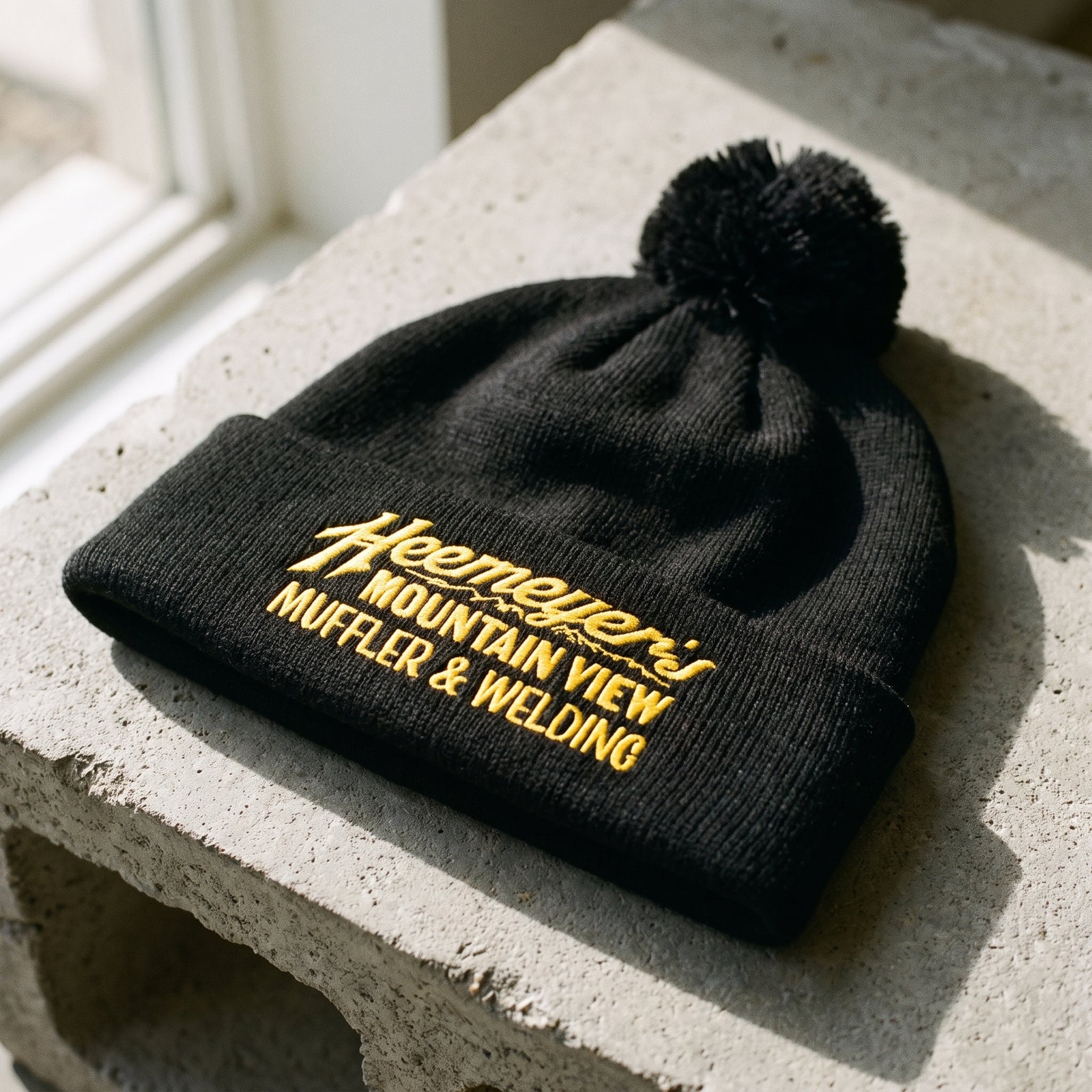Black Heemeyer’s Mountain View Muffler embroidered pom-pom beanie, close-up flat lay on concrete