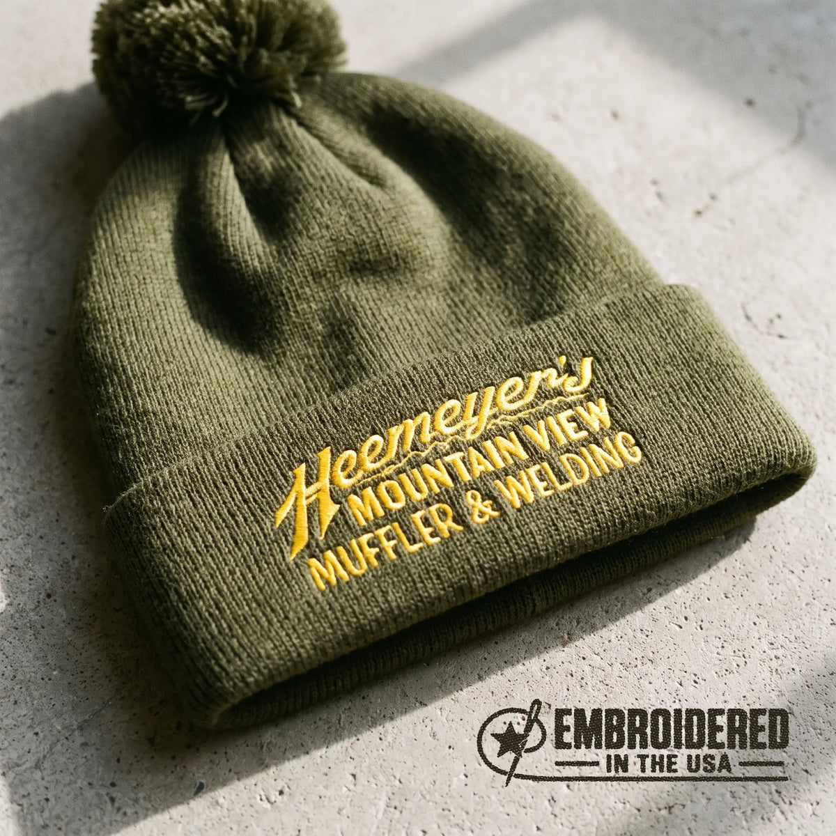 Moss green Heemeyer’s Mountain View Muffler embroidered pom-pom beanie, close-up flat lay on concrete