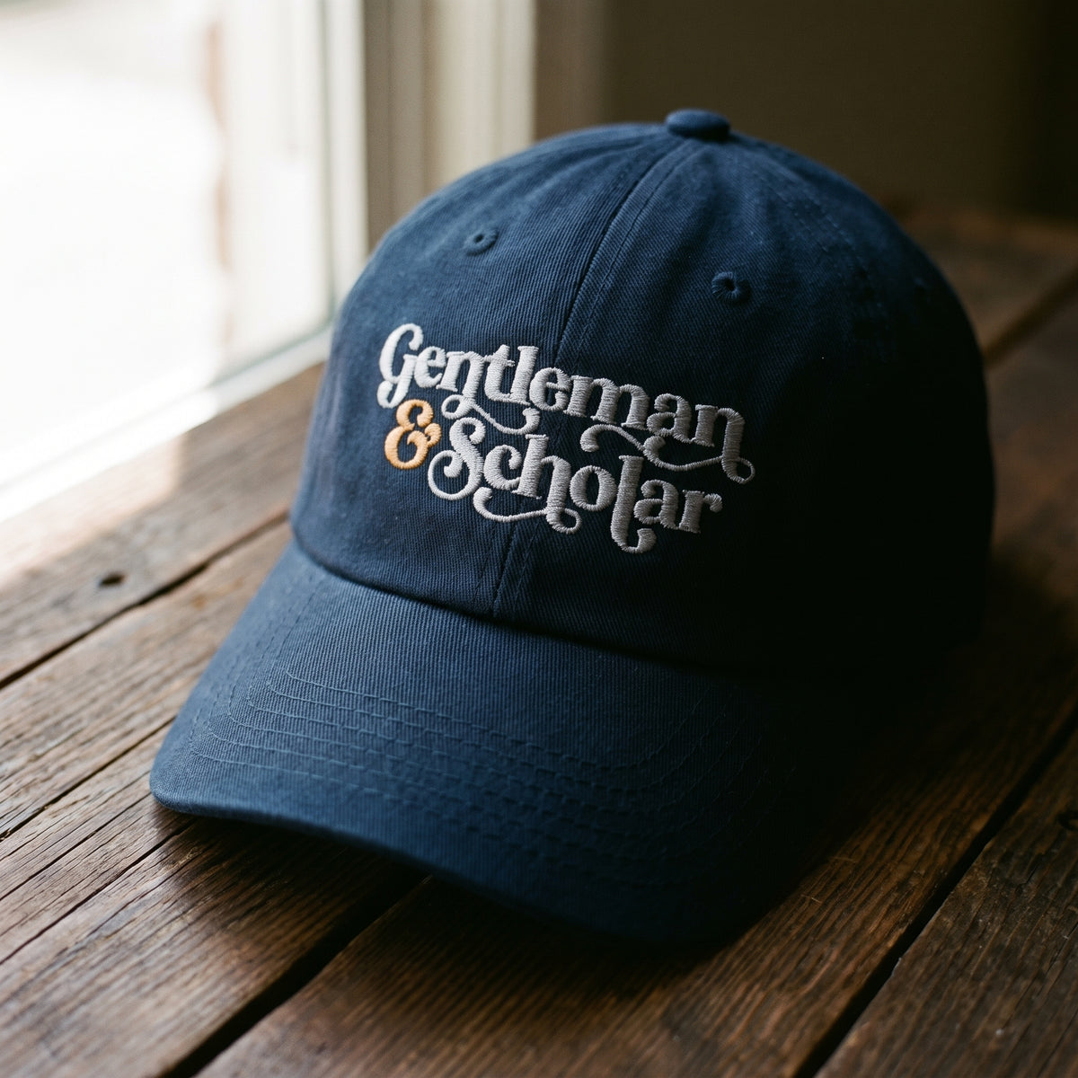 A navy blue baseball cap with 'Gentleman &amp; Scholar' embroidered on the front, resting on a rustic wooden table near a window.