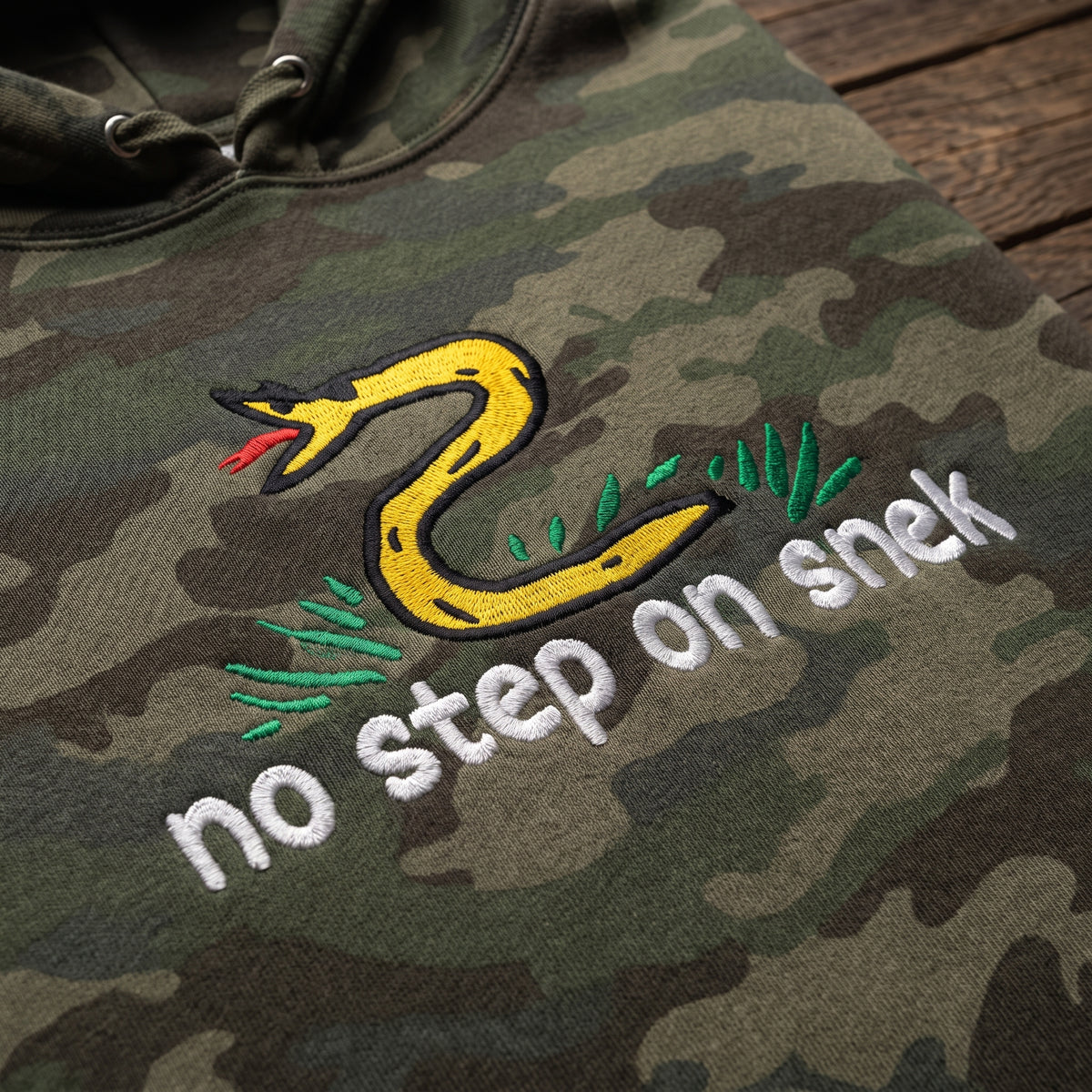 Close-up of No Step on Snek embroidery on Forest Camo hoodie laid on a wood table