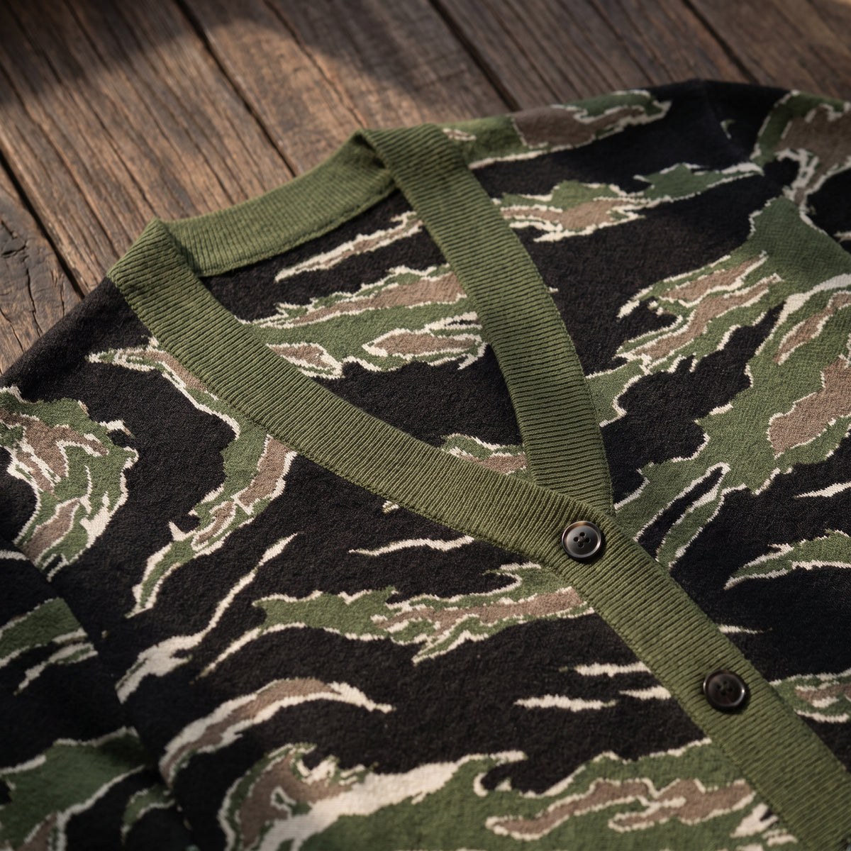 Close-up of a knit cardigan with a tiger-stripe camouflage pattern in olive green, black, and beige, featuring an olive green ribbed V-neck and button placket, laid flat on a rustic wooden surface.