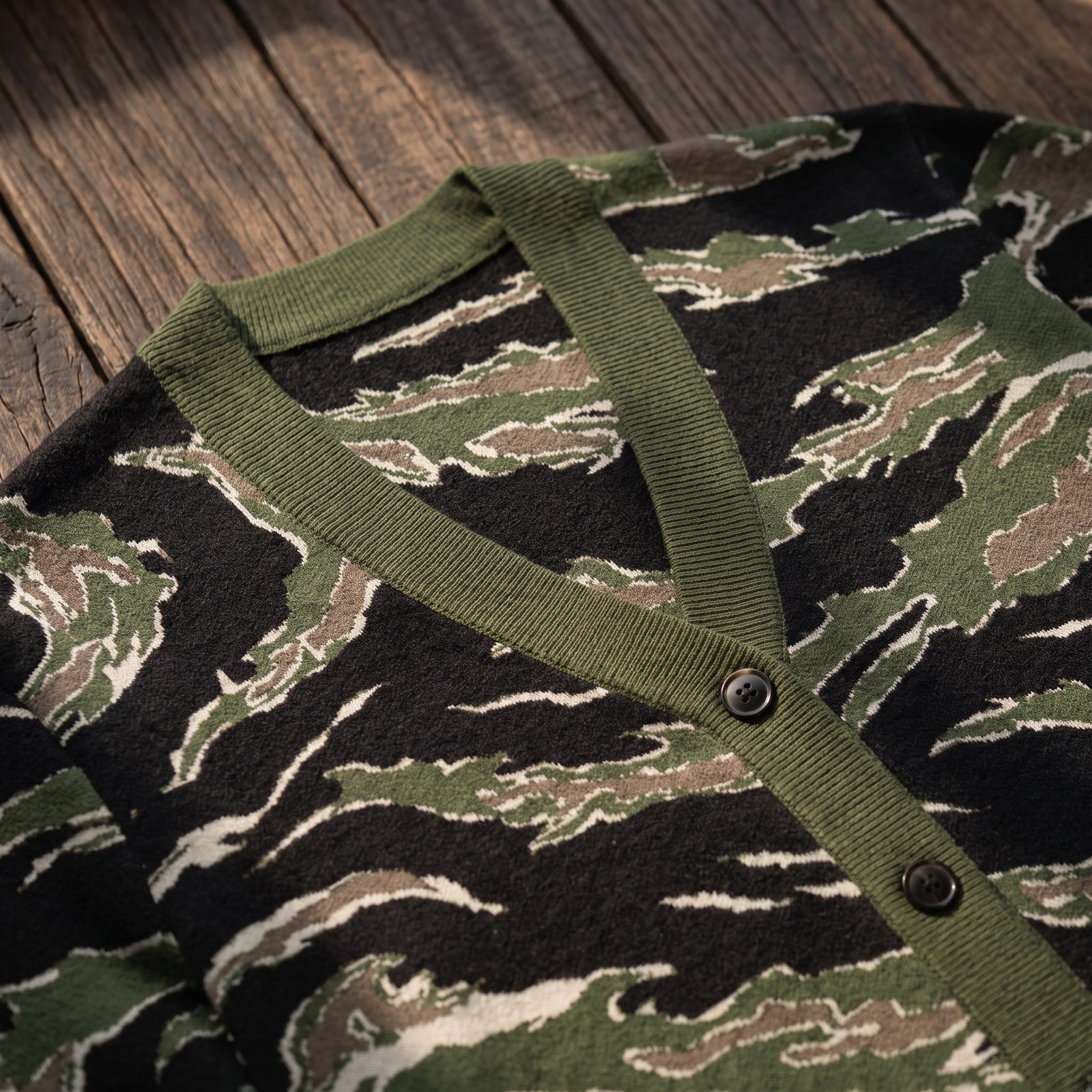 Close-up of a knit cardigan with a tiger-stripe camouflage pattern in olive green, black, and beige, featuring an olive green ribbed V-neck and button placket, laid flat on a rustic wooden surface.