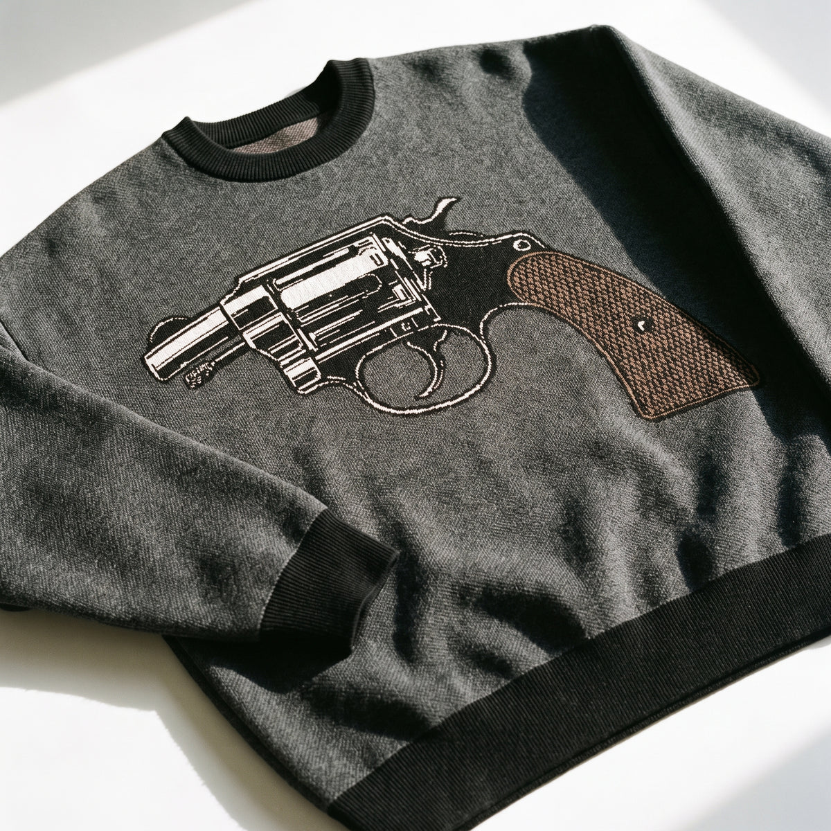 A dark gray crewneck Jacquard-kni sweater featuring a large graphic of a revolver pistol on the front, laid flat on a white surface with strong natural sunlight casting shadows.