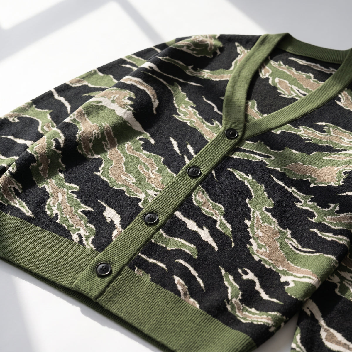 A close-up of a knit cardigan with a tiger-stripe camouflage pattern in olive green, black, and beige, featuring an olive green ribbed V-neck, button placket, and cuffs.