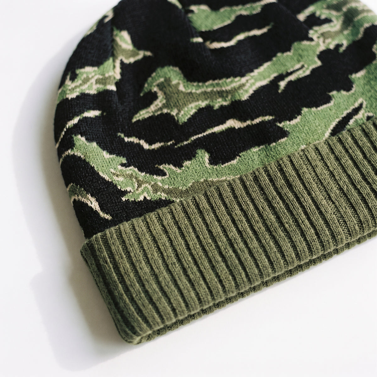 Close-up of a knit beanie with a green and black camouflage pattern and an olive green ribbed cuff, photographed on a white background.