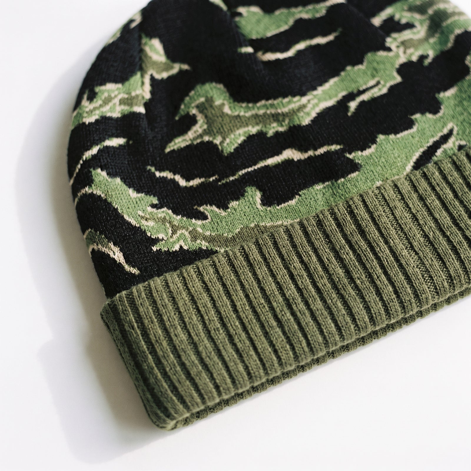 Close-up of a knit beanie with a green and black camouflage pattern and an olive green ribbed cuff, photographed on a white background.