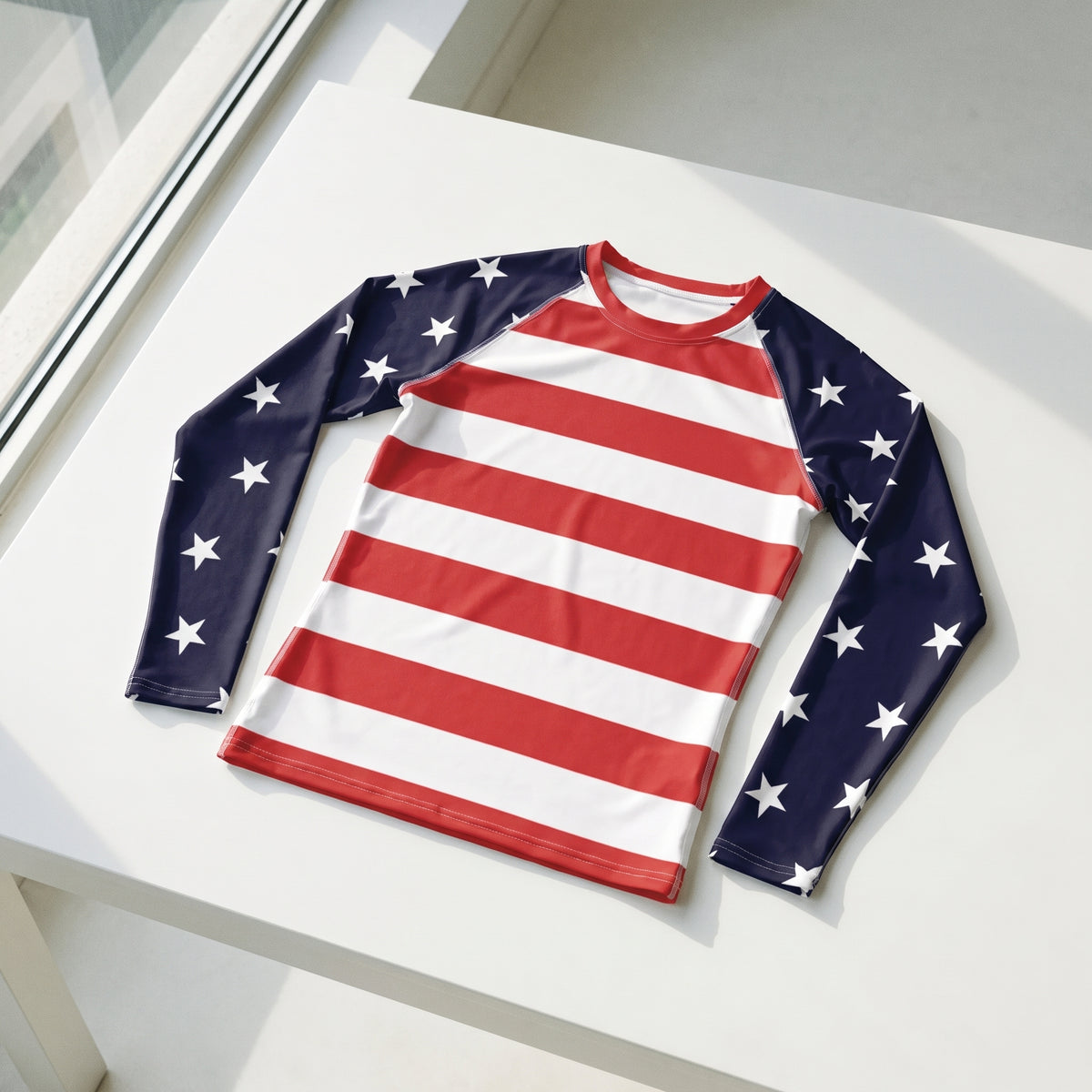 A long-sleeve athletic rash guard with an American flag design, featuring red and white horizontal stripes on the torso and navy blue sleeves with white stars, displayed on a white table.