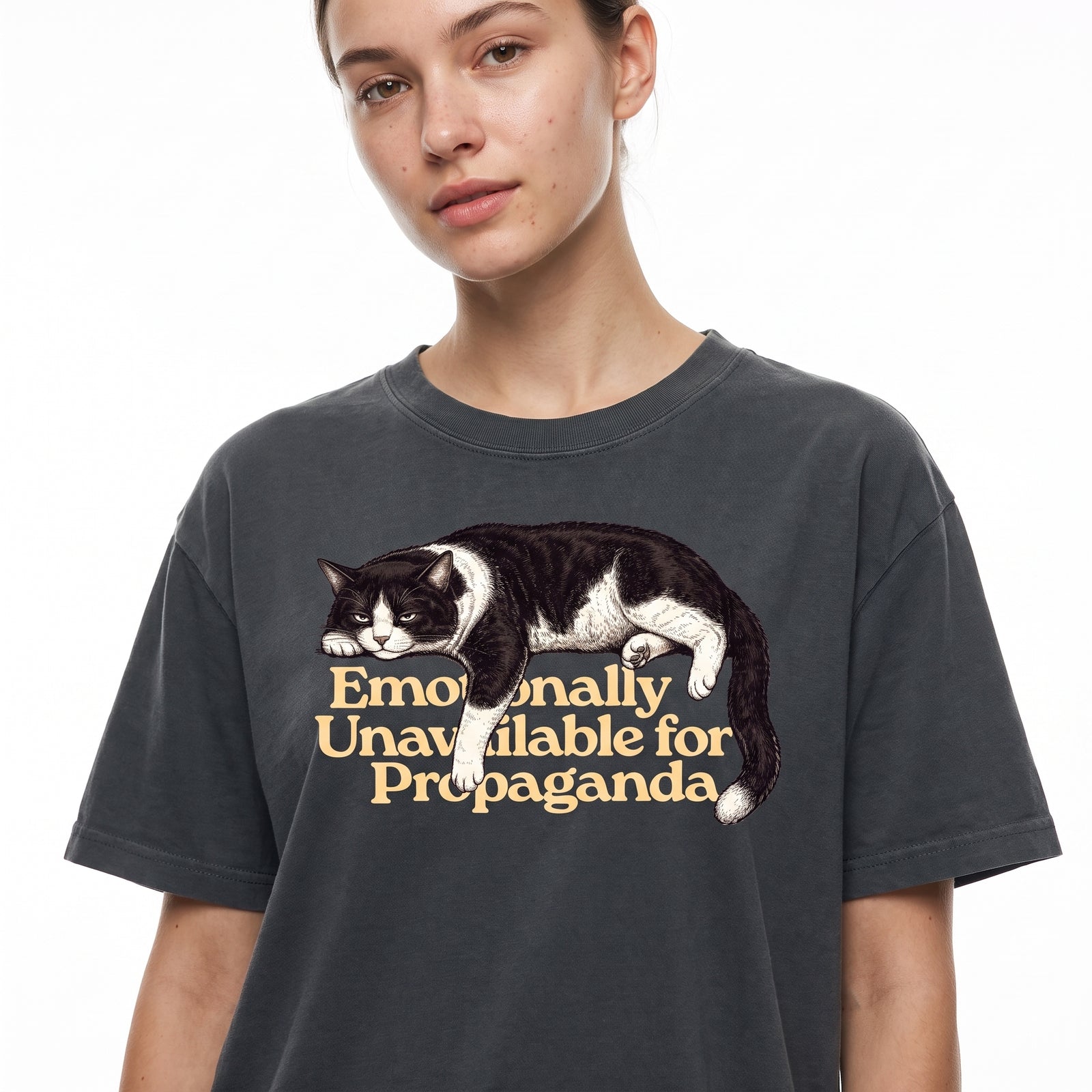 A woman wearing a dark charcoal gray t-shirt with a graphic of a black and white cat lying down next to the text 'Emotionally Unavailable for Propaganda' in a tan serif font, set against a plain white background.