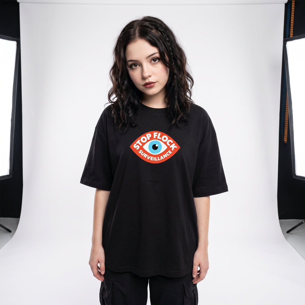 A woman modeling an oversized black t-shirt with a 'STOP FLOCK SURVEILLANCE' graphic on the chest, set against a white studio backdrop with softbox lights visible on the sides.