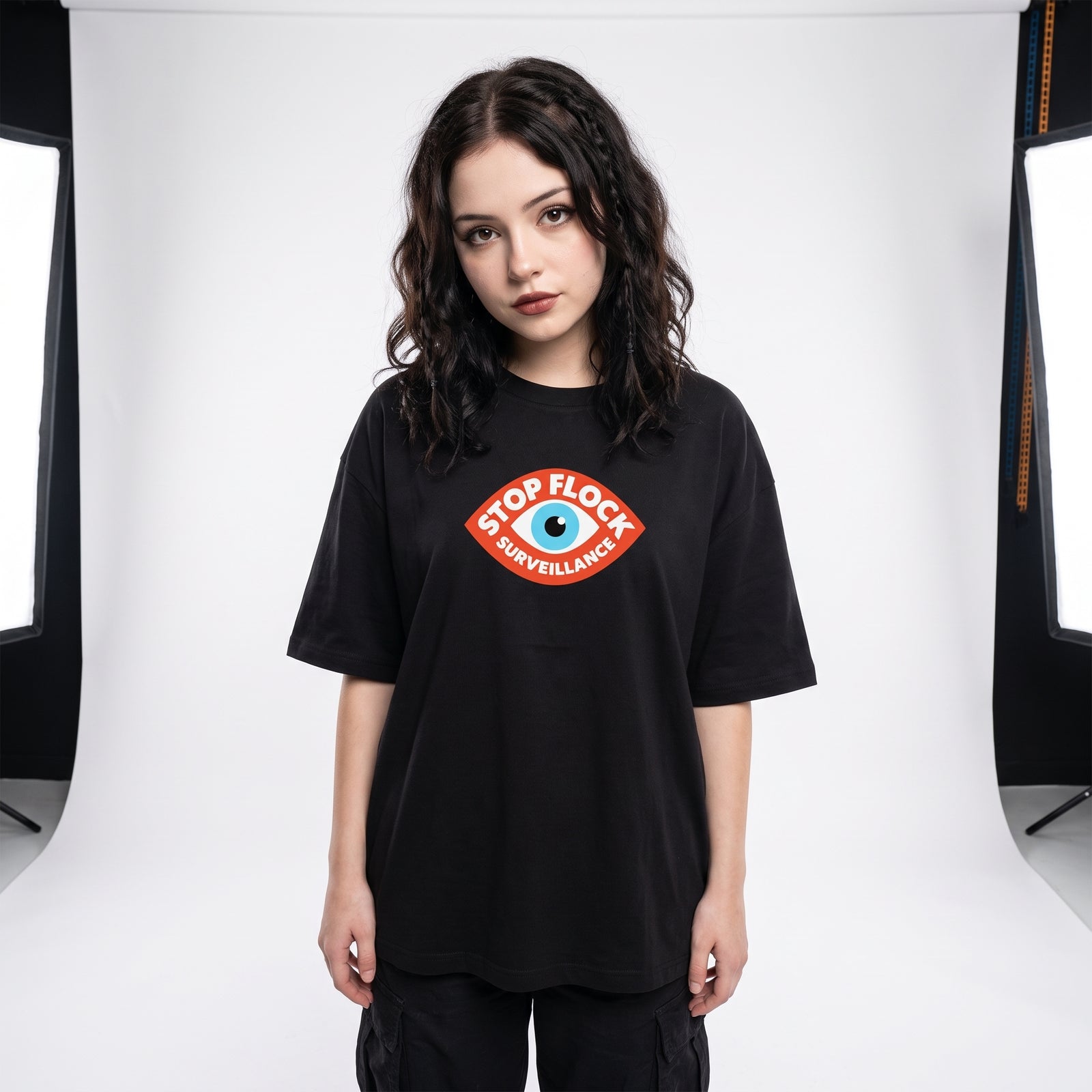 A woman modeling an oversized black t-shirt with a 'STOP FLOCK SURVEILLANCE' graphic on the chest, set against a white studio backdrop with softbox lights visible on the sides.